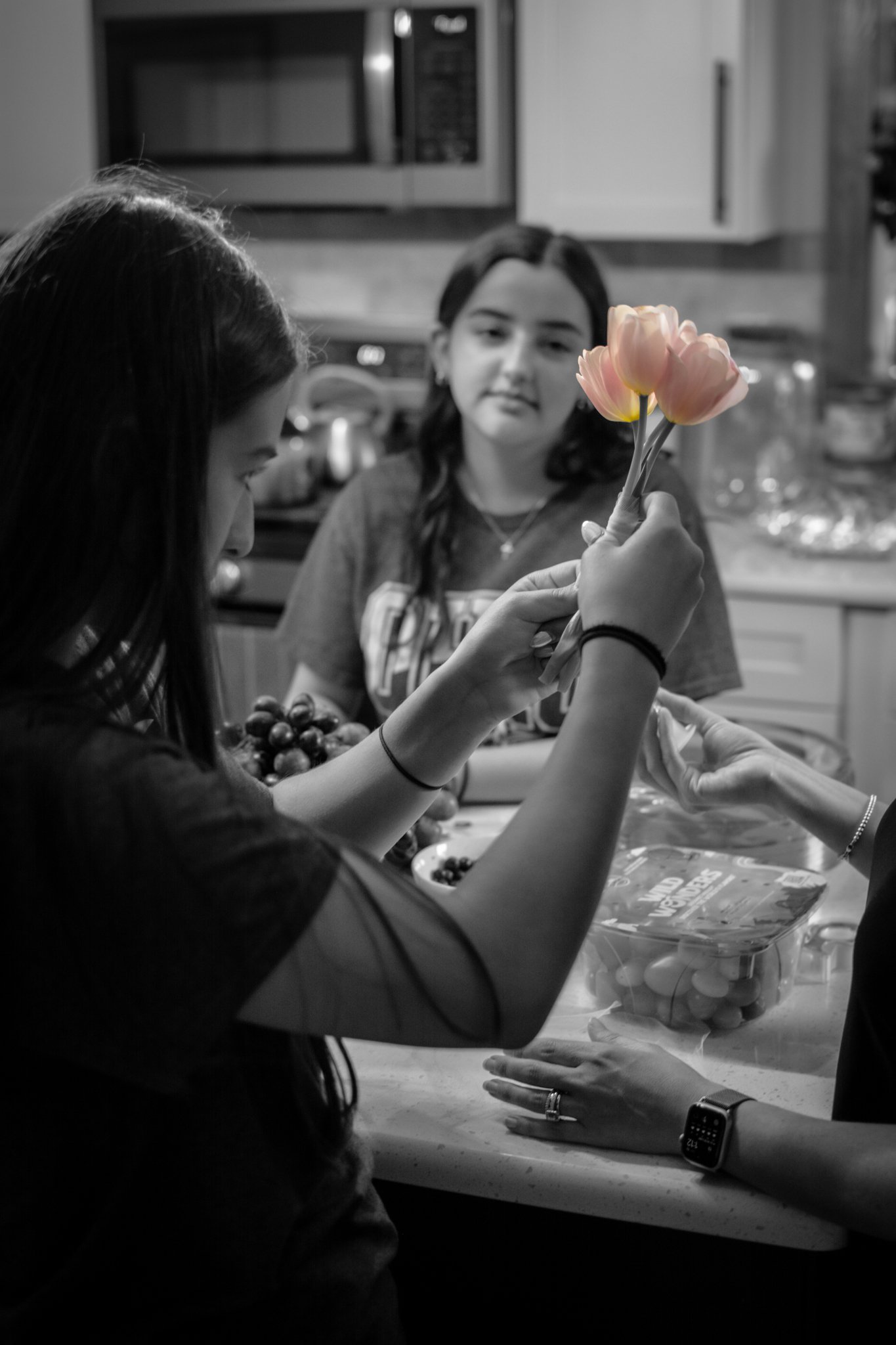 Two young girls are in a kitchen, one is holding a handful of grapes, and the other is holding a bouquet of pink tulips. The scene is captured in black and white, with the tulips in color.