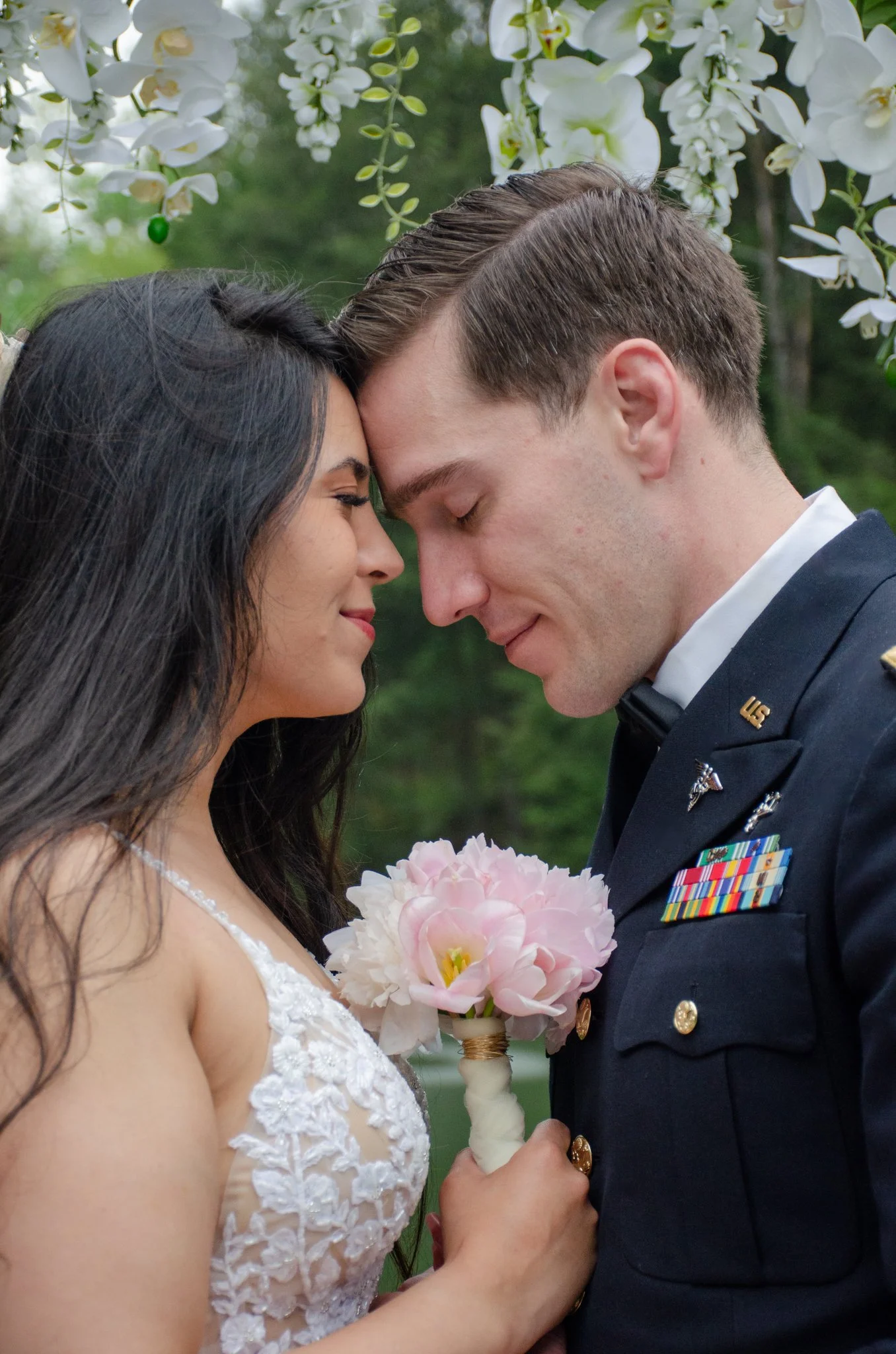 A bride and groom in a close, intimate moment with foreheads touching, holding a pink flower bouquet, outdoors with white flowers hanging above them.