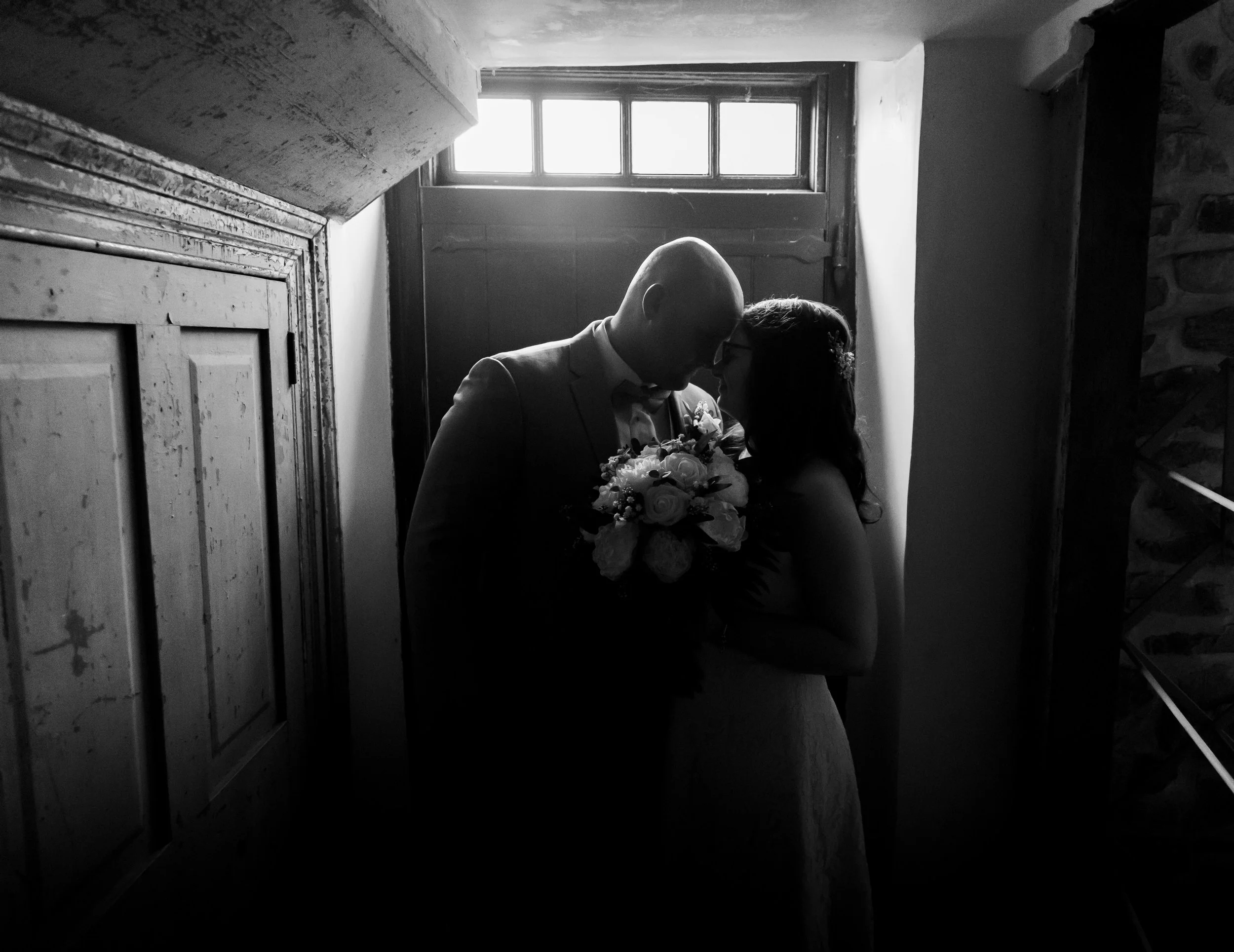 Black and white photo of a bride and groom standing close indoors, with only their silhouettes visible as their foreheads touch. The groom holds a bouquet of flowers, and the background shows a small window above a door.