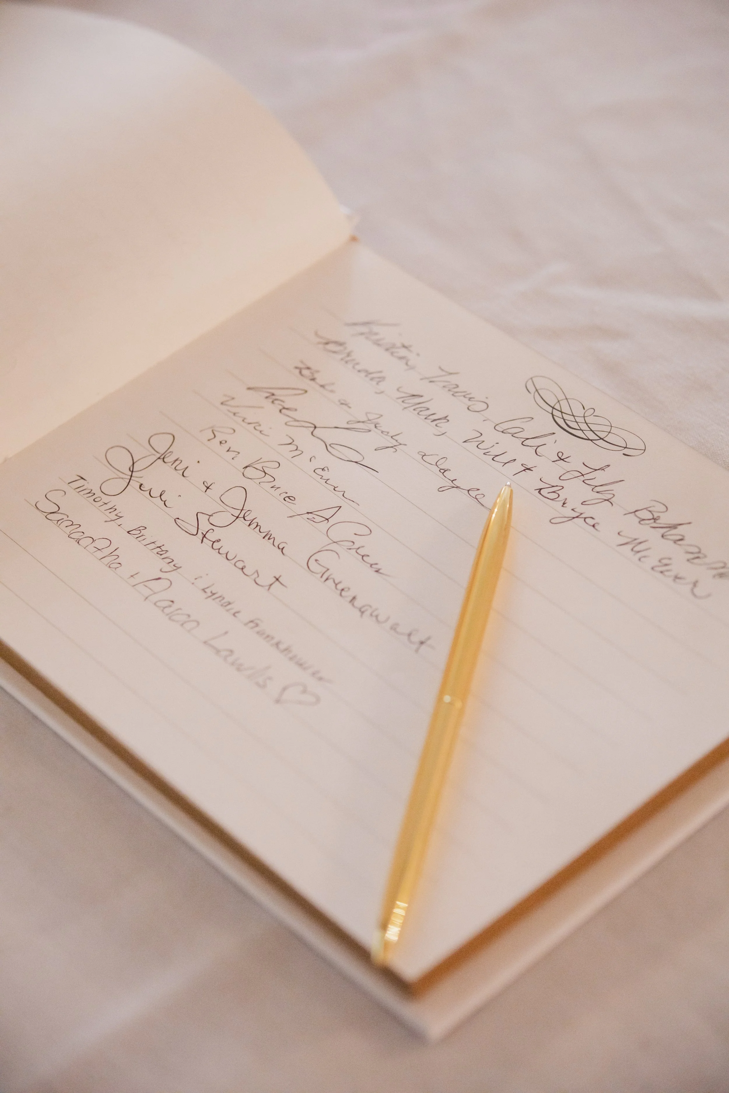 A guestbook with handwritten signatures and messages, accompanied by a gold pen, on a white tablecloth at a wedding or special event.