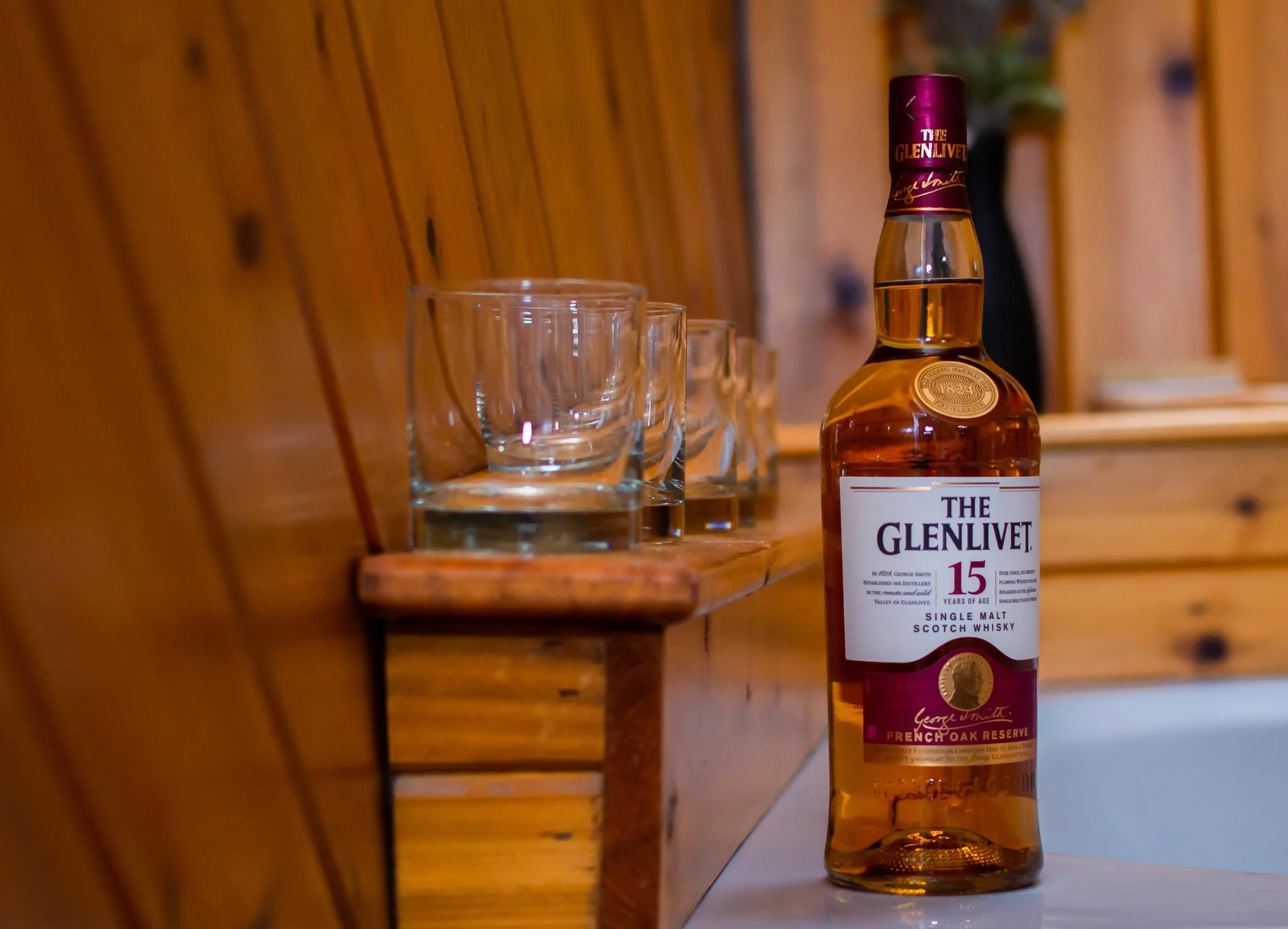 A bottle of Glenlivet 15-year-old single malt Scotch whisky placed next to four glasses of whisky on a wooden shelf.