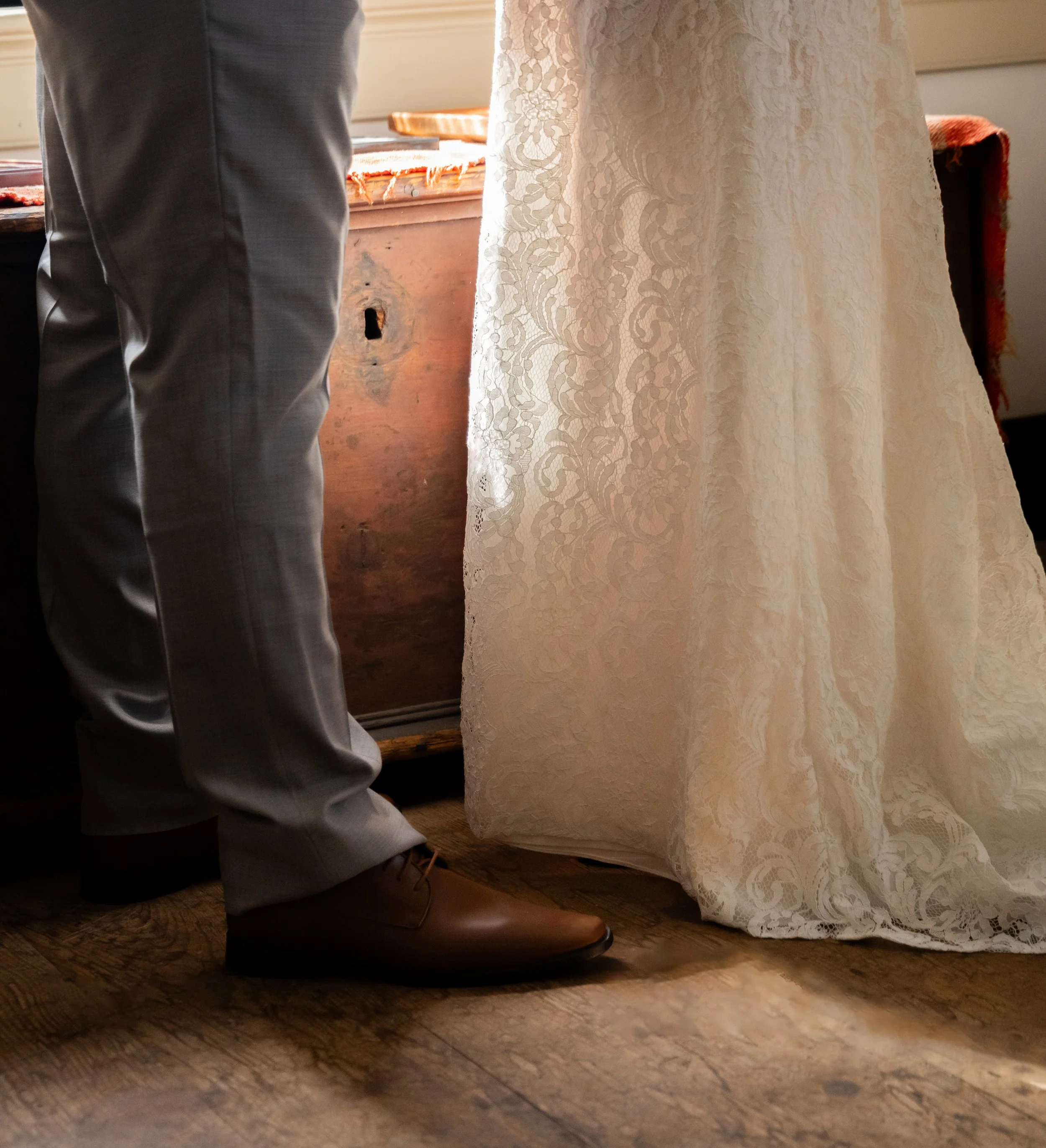 Partially visible person in gray pants and brown shoes standing beside a table with a lace wedding dress hanging nearby.