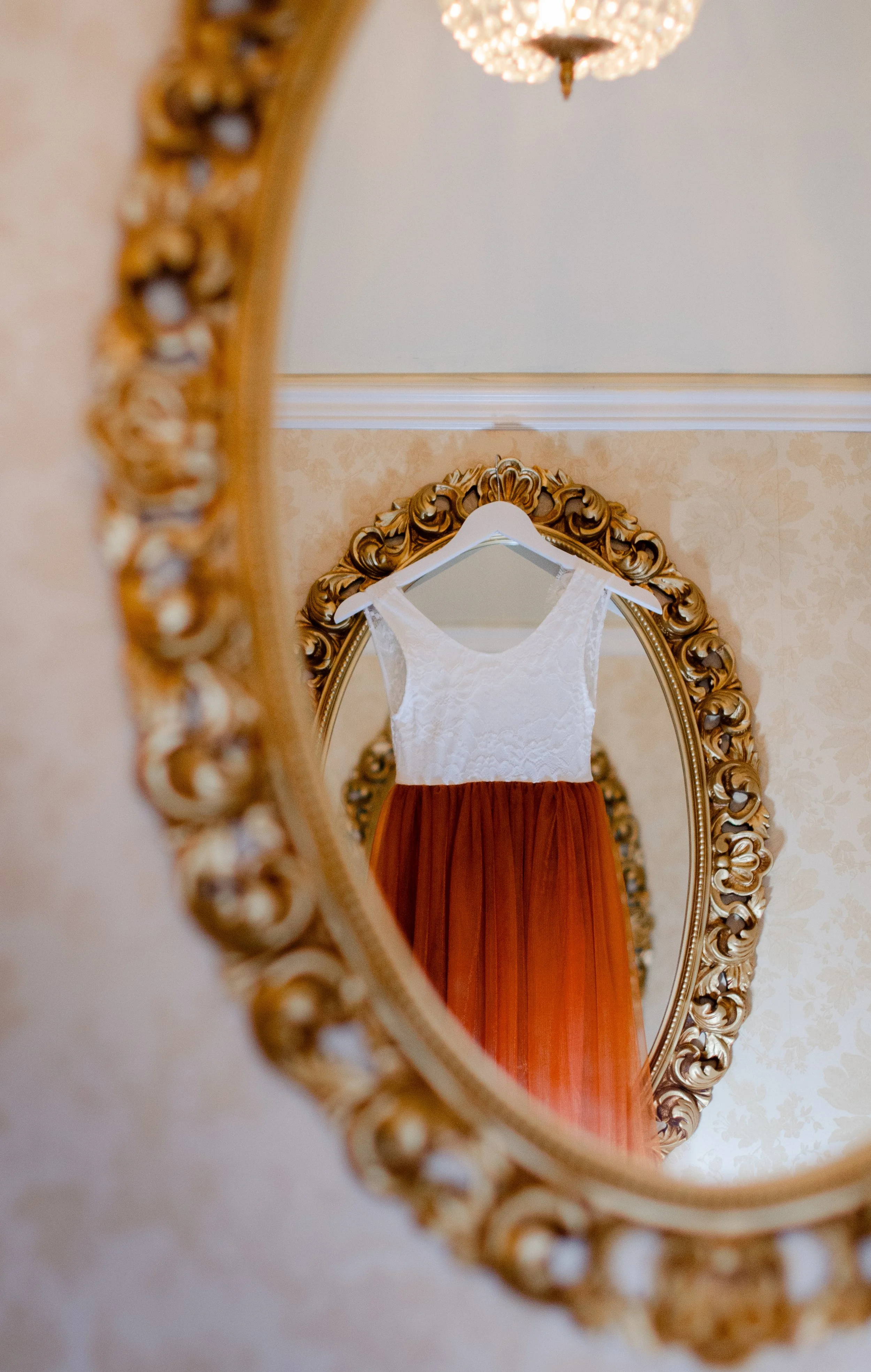 A wedding dress with a white lace top and a rust-colored pleated skirt hanging on a white hanger, reflected in an ornate gold-trimmed mirror.