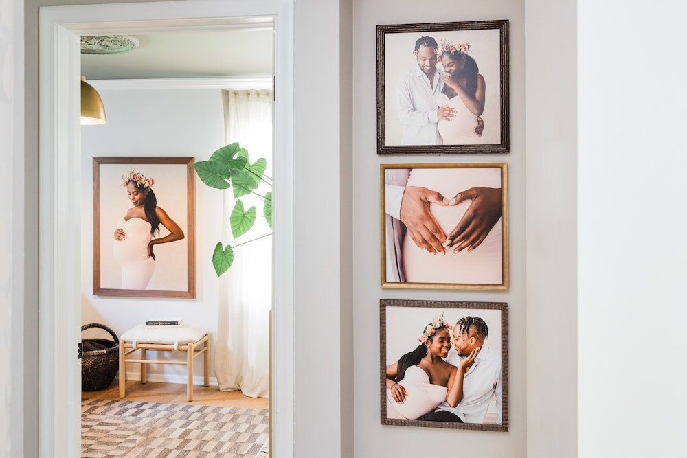 A cozy corner of a home with four framed family and couple photographs on the wall. The wall features images of a pregnant woman, a couple making a heart shape with their hands, and a couple smiling and embracing. A large green plant with heart-shaped leaves is present, and a bench and a basket are visible in the room beyond.