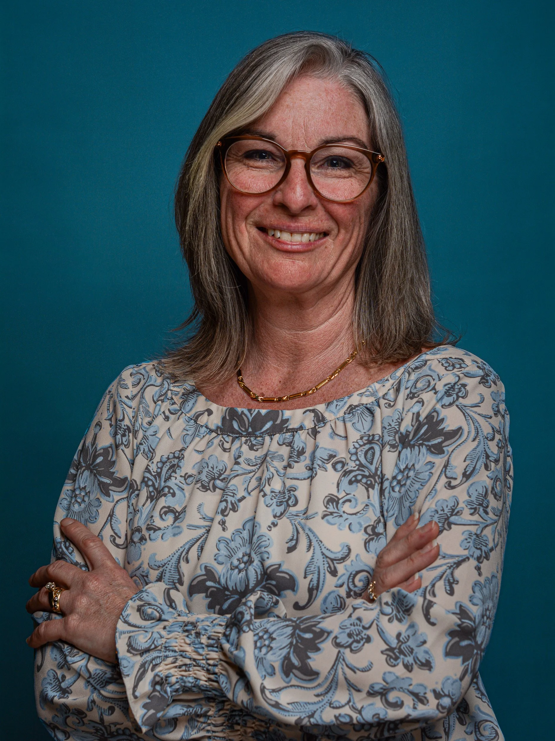 A woman with shoulder-length gray hair and glasses, smiling, wearing a floral blouse with blue and white pattern, a gold necklace, and rings, standing with arms crossed against a blue background.