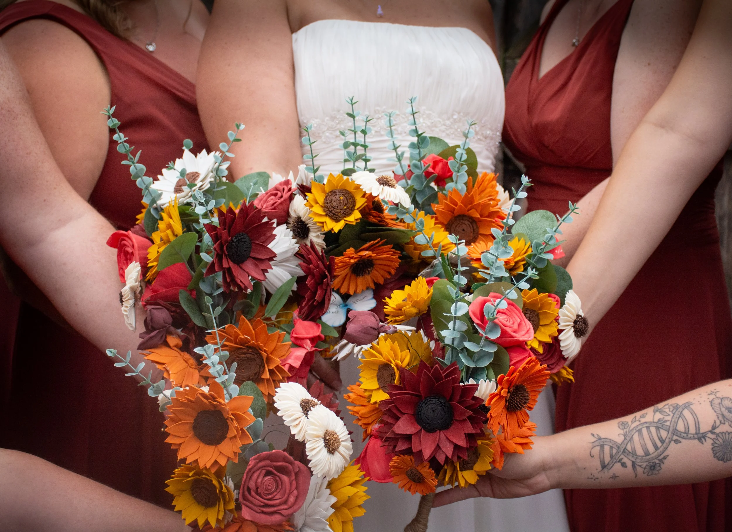 A group of women in burgundy dresses holding a vibrant bouquet of artificial flowers, including sunflowers, daisies, roses, and eucalyptus, at a wedding or celebration.