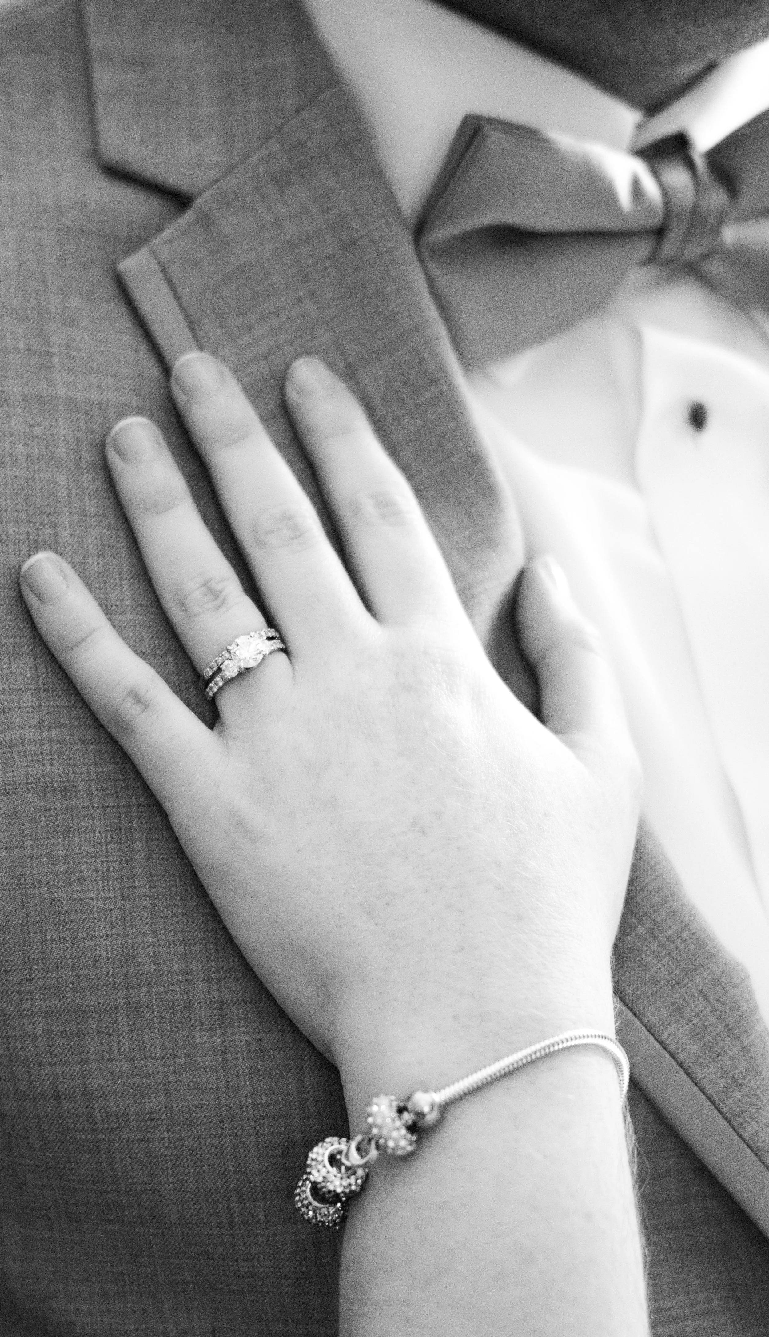 A woman's hand wearing a diamond ring and a bracelet rests on a man's suit jacket, indicating a wedding or engagement.