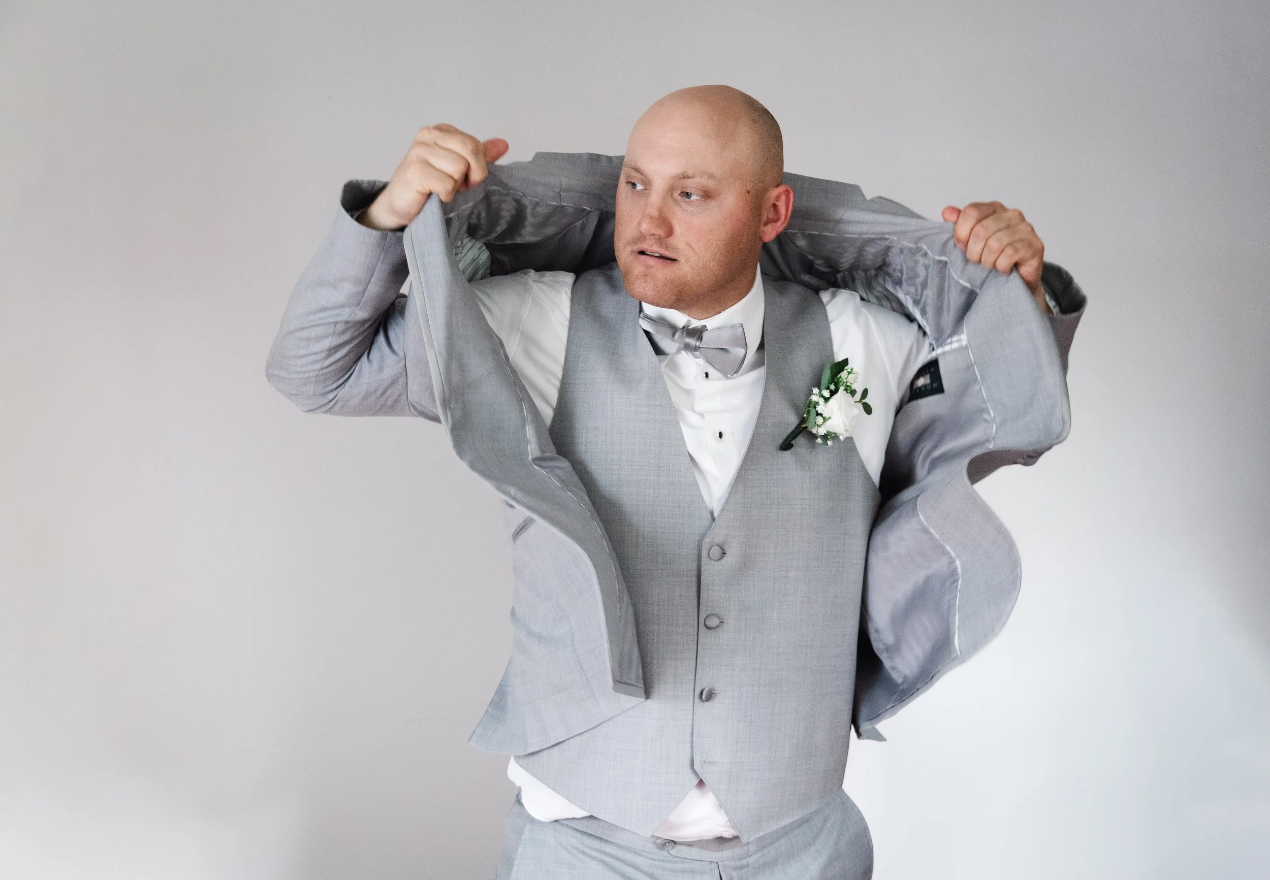 A man in a light gray suit and bow tie adjusting his suit jacket against a plain gray background.