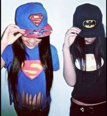 Two girls wearing superhero-themed caps, one with a Superman logo and the other with a Batman logo, smiling and looking down.