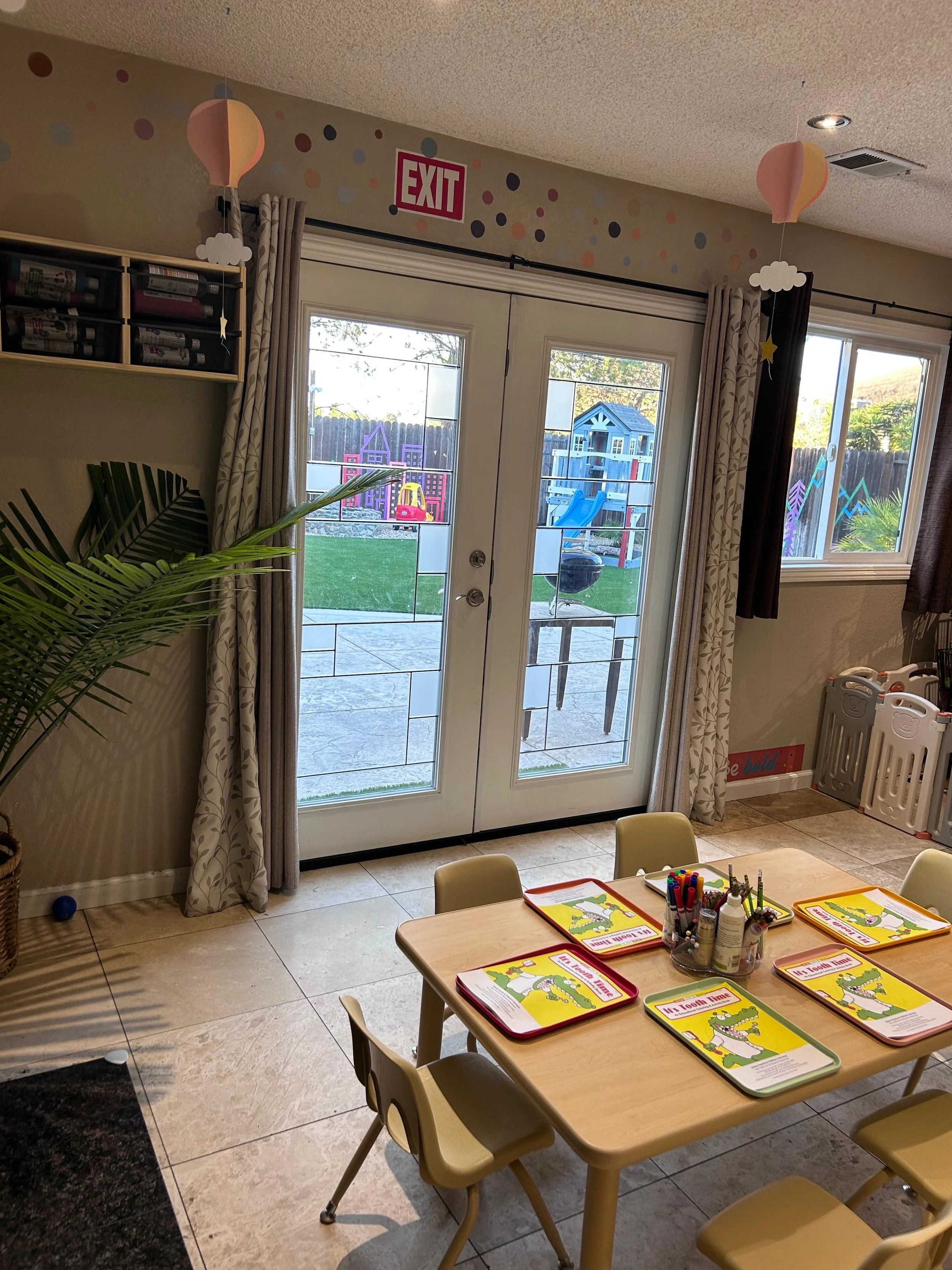Indoor children's classroom with small chairs around a table, papers, and art supplies, with child-friendly decor and a view of a backyard with a playset outside.