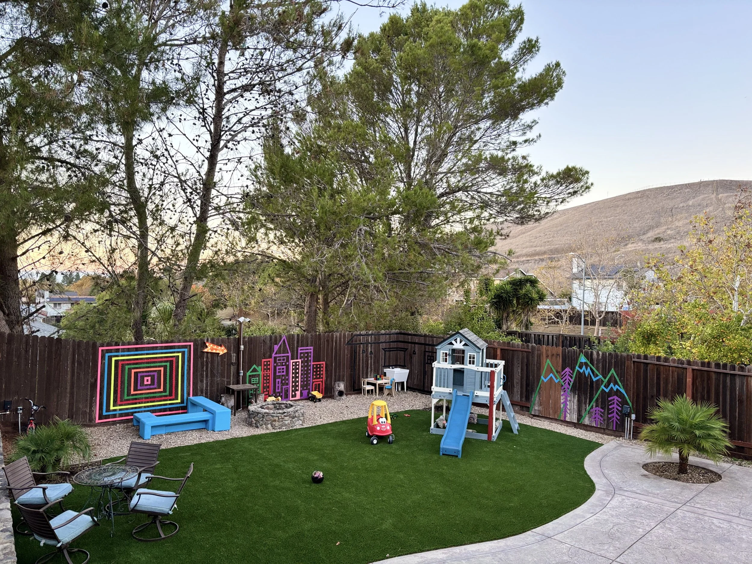 Colorful fenced backyard with a playhouse, slide, toys, and outdoor furniture, decorated with painted wall art of buildings and mountains, surrounded by trees and a mountain hill in the background.