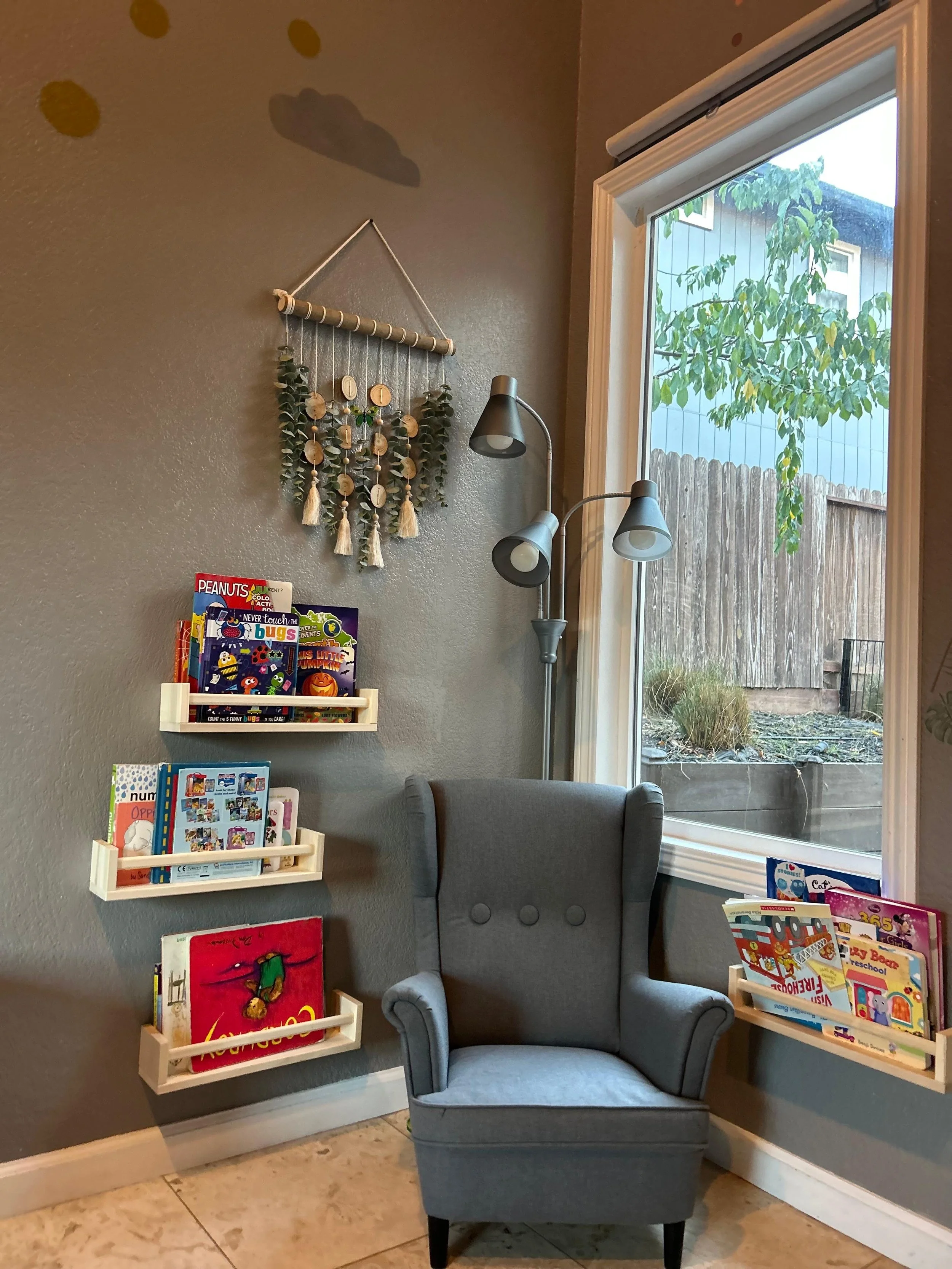 A reading nook with a gray armchair, three wall-mounted book shelves filled with children's books, a wall hanging made of hanging wood circles and greenery, and a ground lamp with three adjustable heads. A large window shows an outdoor garden with a 