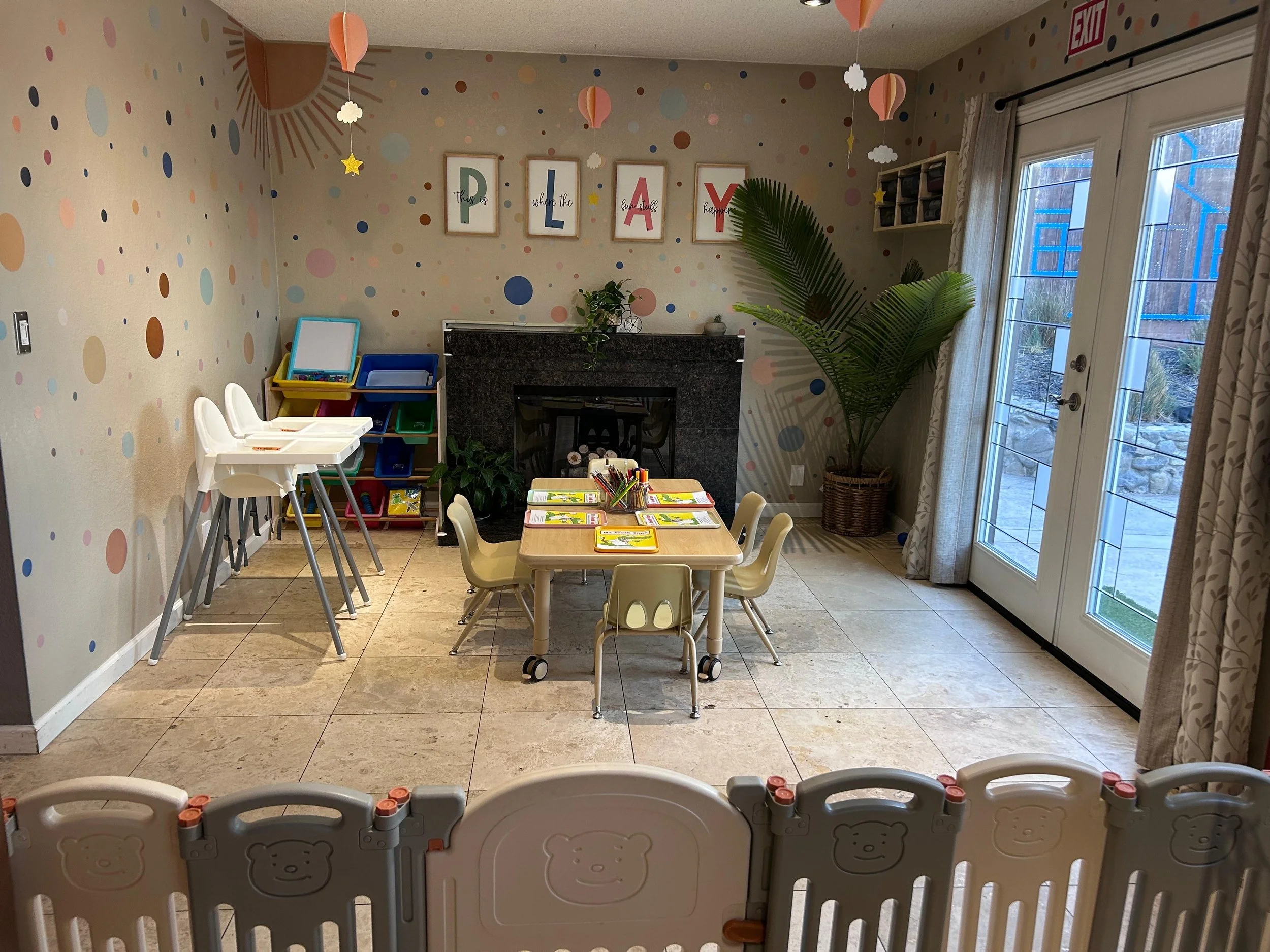 A cozy children's playroom with a polka dot patterned wall, a fireplace with a plant on top, and a large potted palm in the corner. There are four child-sized chairs around a small table with art supplies, and a white safety gate borders the foregrou
