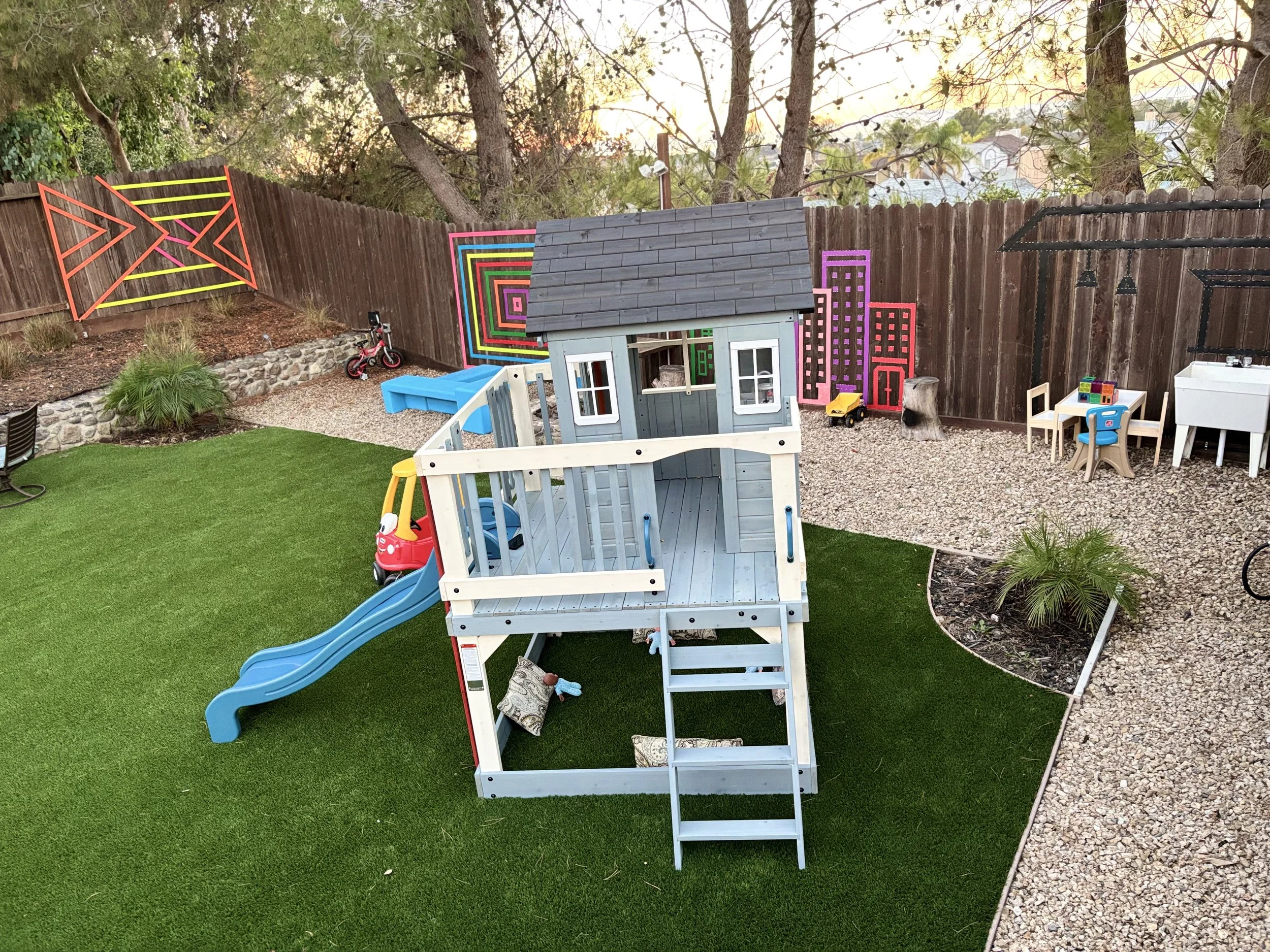 Backyard with a playhouse, slide, and children's toys. Artwork on the fence, including colorful geometric and cityscape murals.