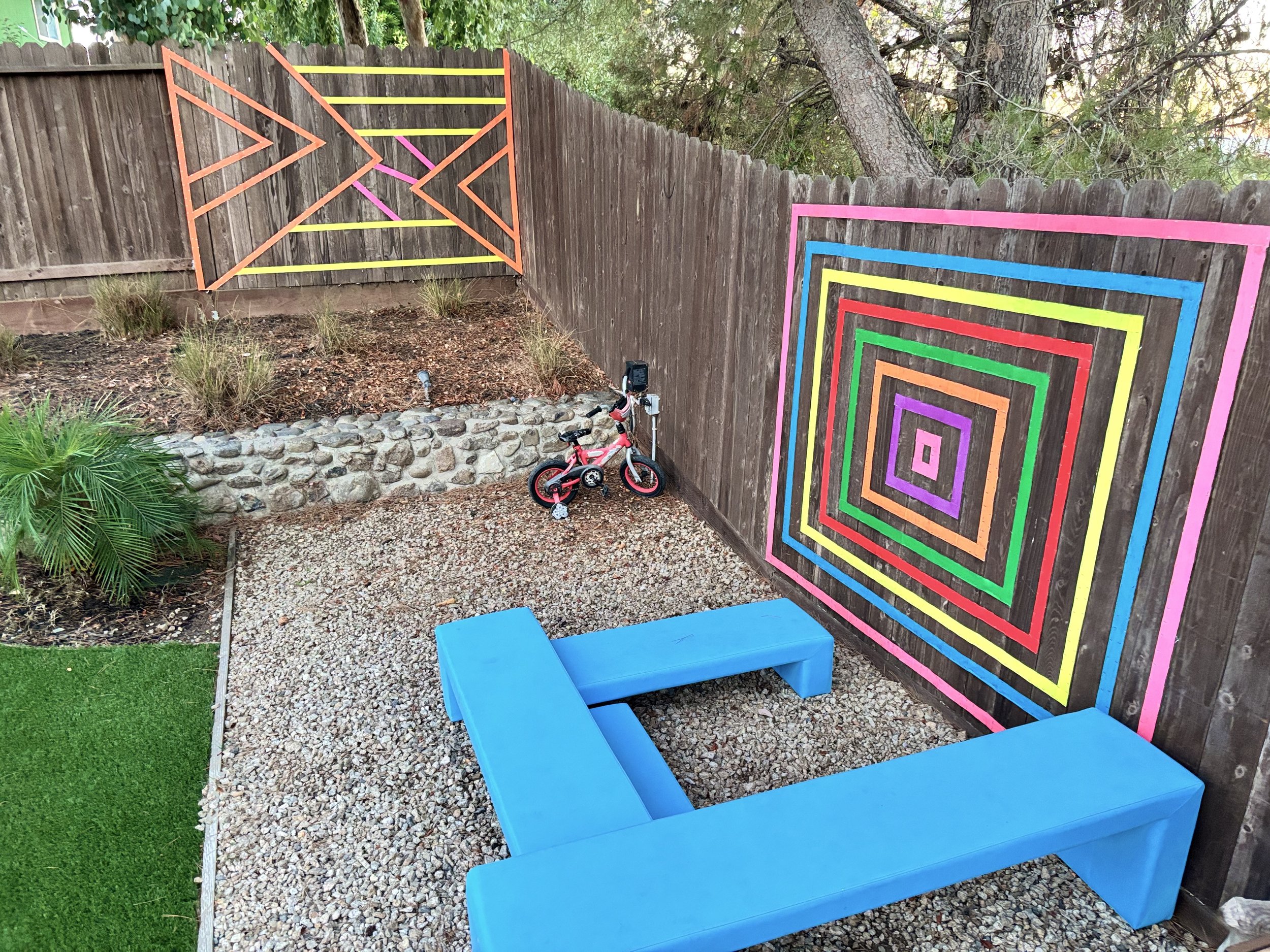 Colored geometric murals on a backyard fence, a blue bench, a small pink bicycle, and a gravel ground with some greenery.