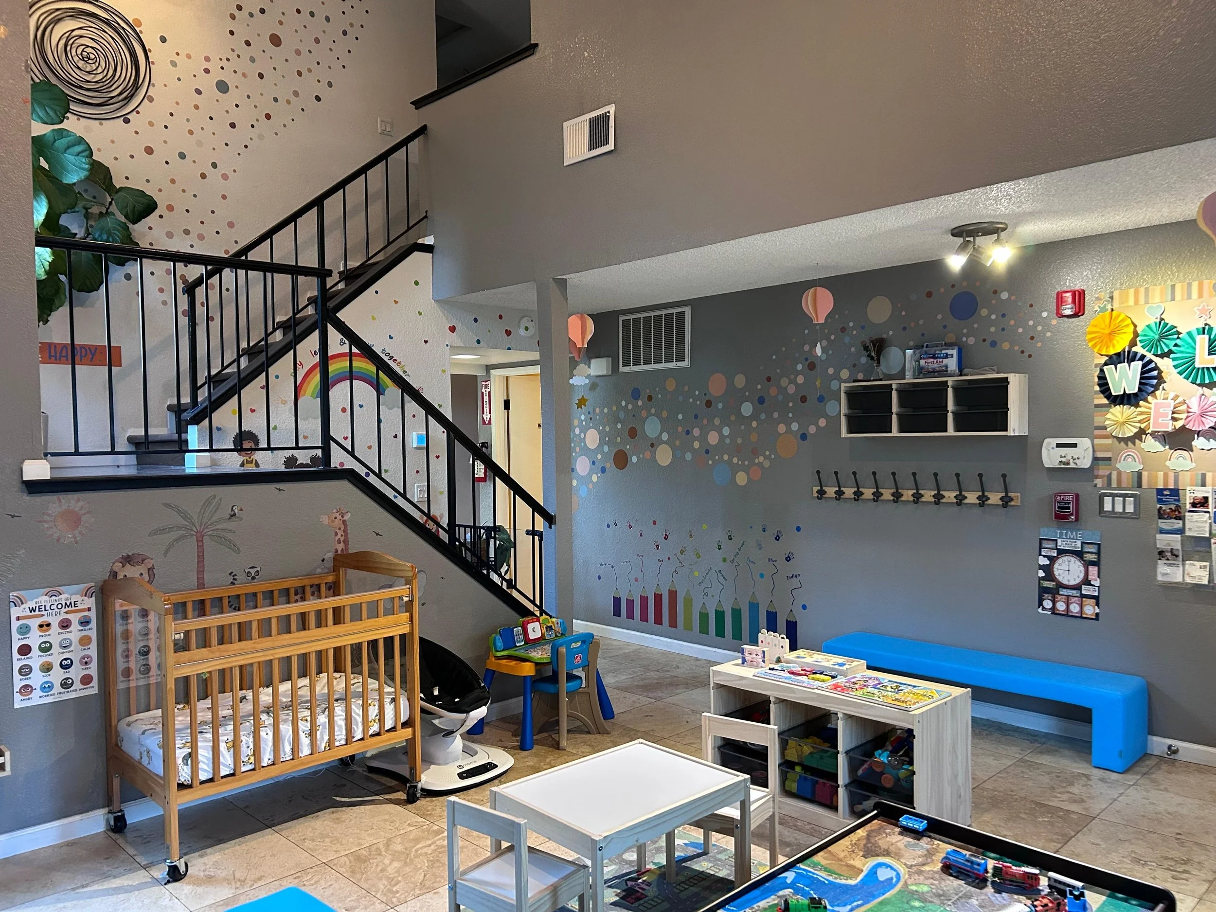 Indoor children's play and learning area with toys, a crib, colorful wall decorations including rainbows, balloons, and a birthday cake mural, small tables and chairs, and various educational materials.