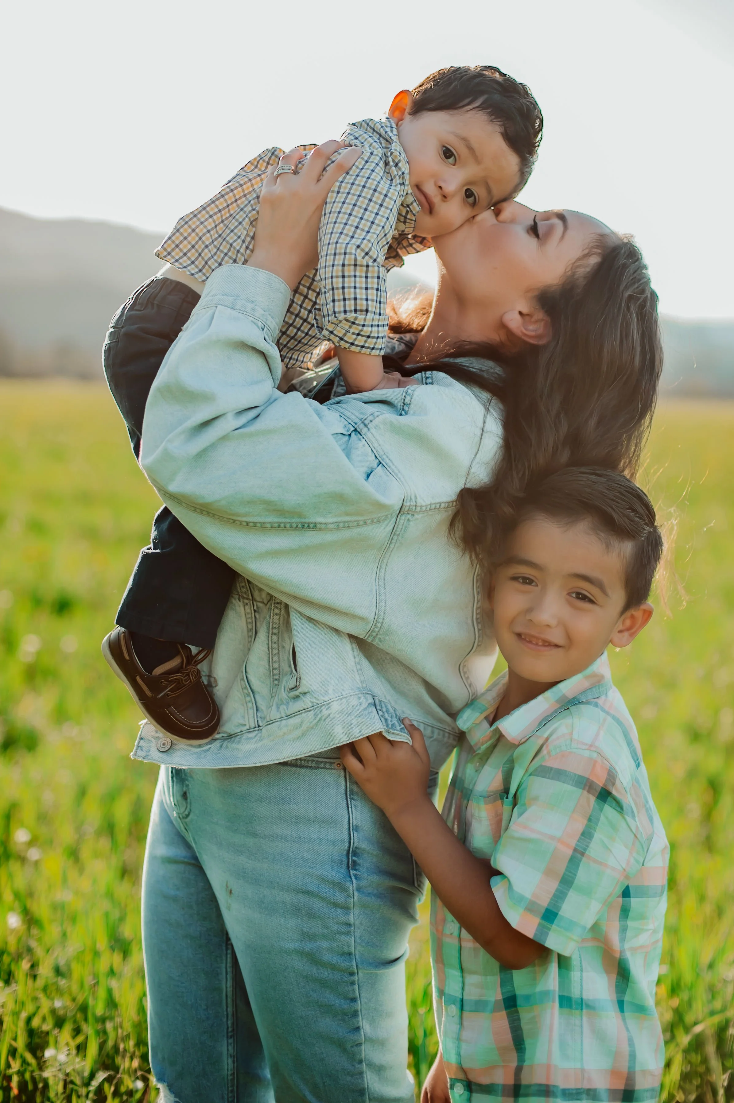 A woman holding a young boy on her shoulder, kissing his forehead, while another boy stands next to her hugging her waist, in a grassy outdoor setting during sunset.
