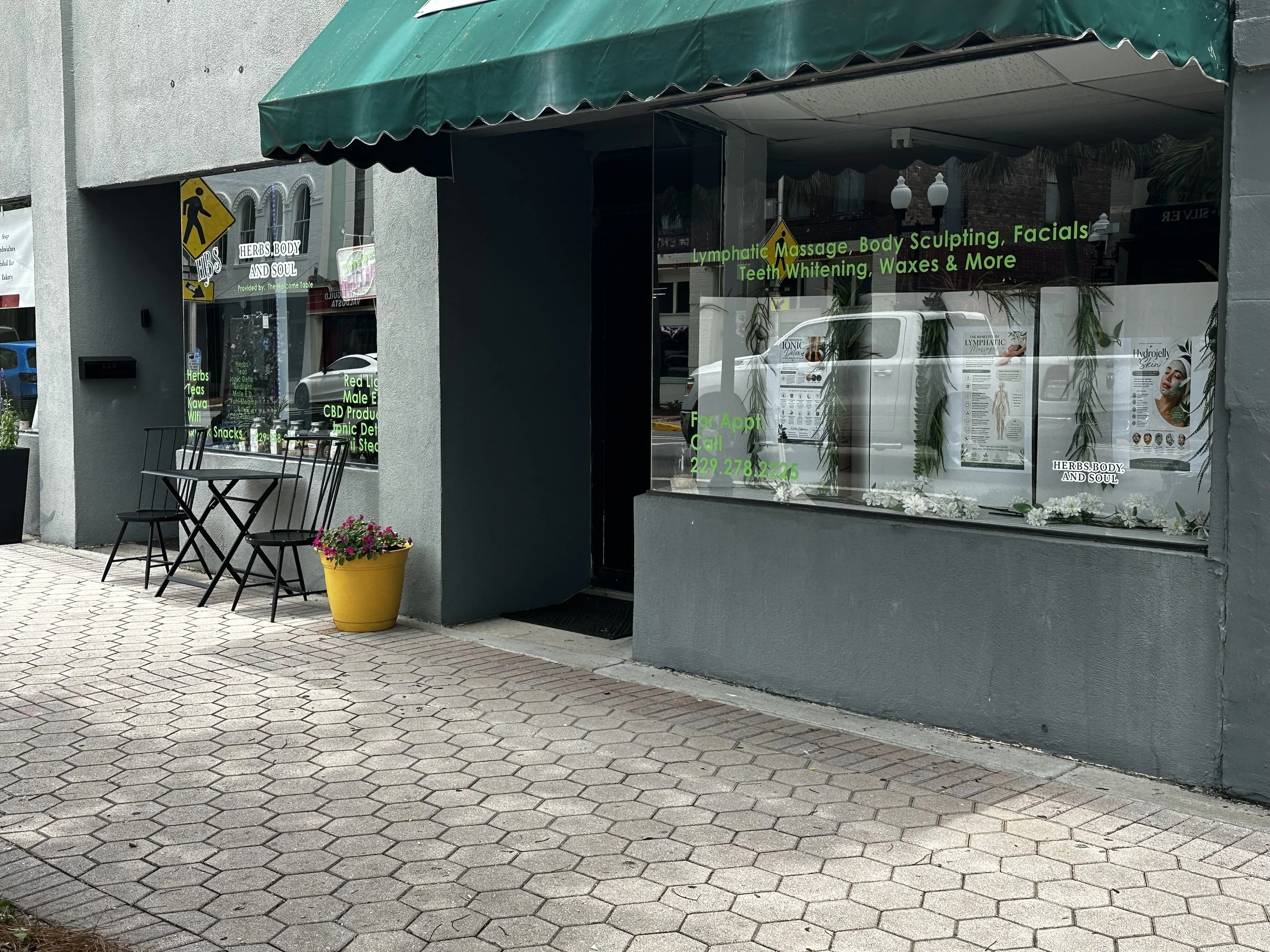 The storefront of a wellness clinic named 'Herbs, Body and Soul' with green neon lettering, a green awning, black chairs and a yellow flowerpot with pink flowers outside. The window displays services such as lymphatic massage, body sculpting, facials, teeth whitening, and waxes, along with a sign for appointments and a phone number.
