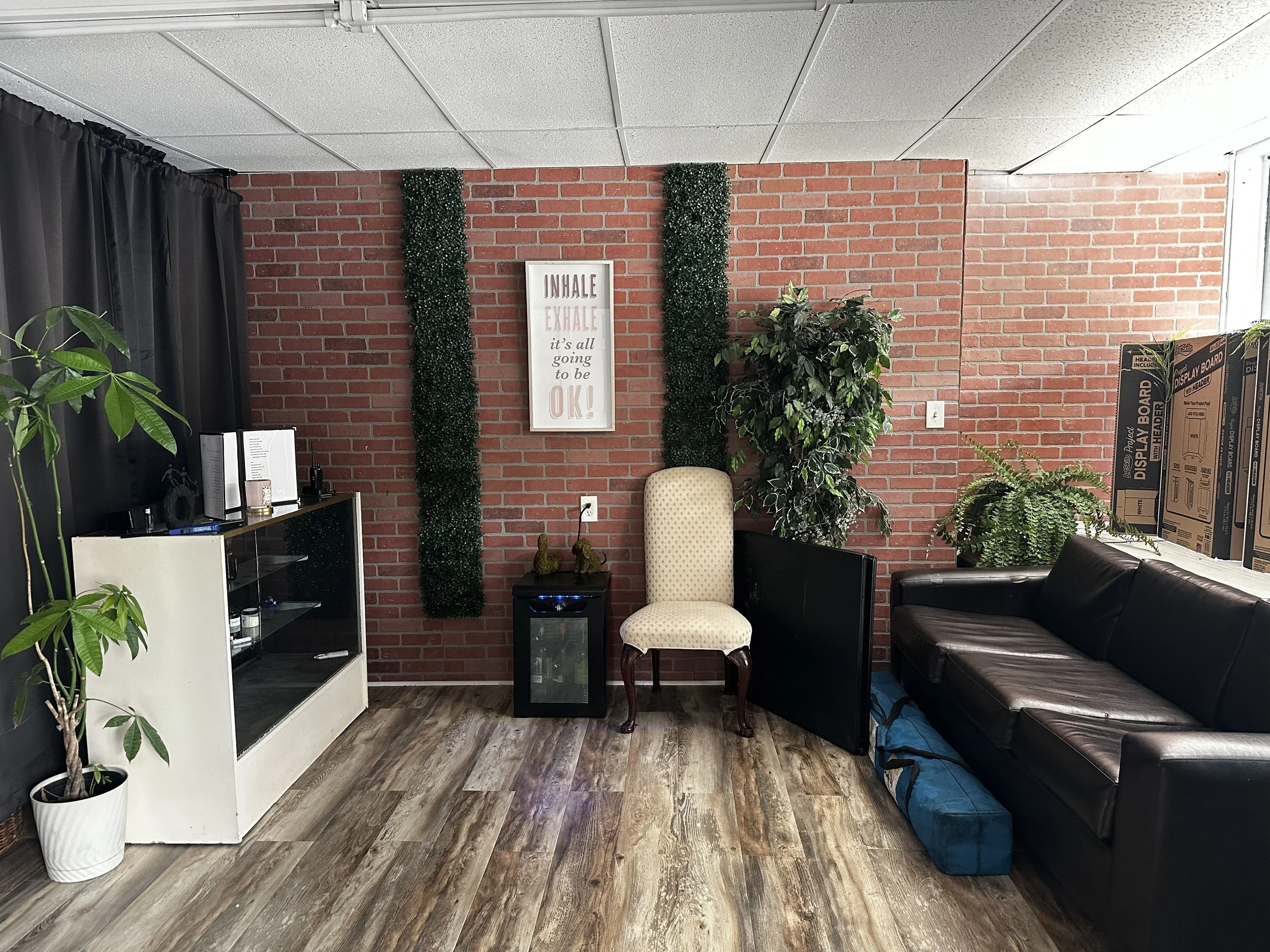 Indoor waiting or lounge area with brick wall backdrop, plants, a framed quote poster, chairs, a black leather couch, and stacked boxes.