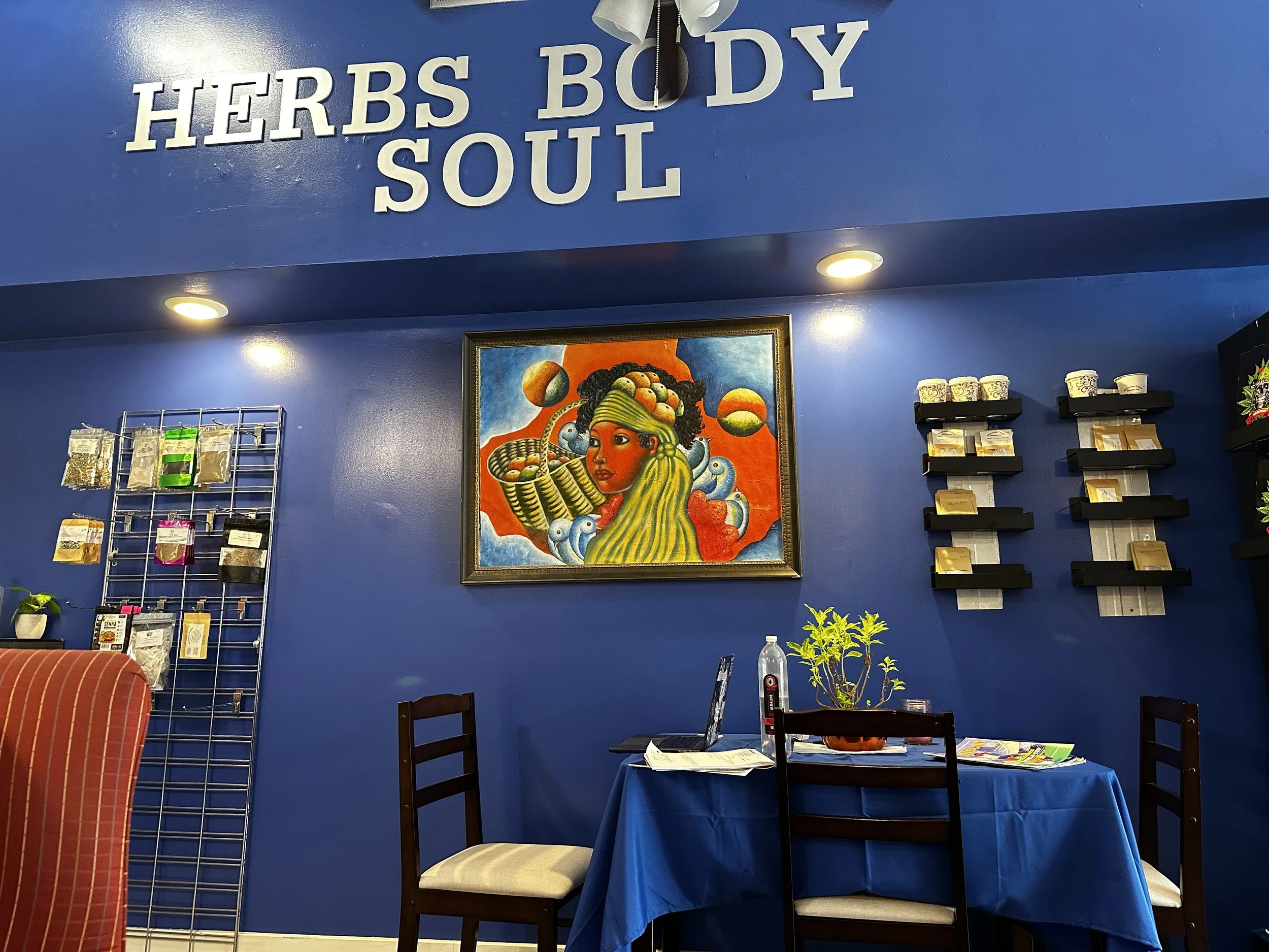 Interior of a store or cafe with a blue wall displaying large white letters spelling "HERBS BODY SOUL." There is a framed colorful artwork of a woman with dark curly hair and a basket of fruits on her head. Shelves with various products, a table with a laptop, papers, and a small plant in a pot are visible.