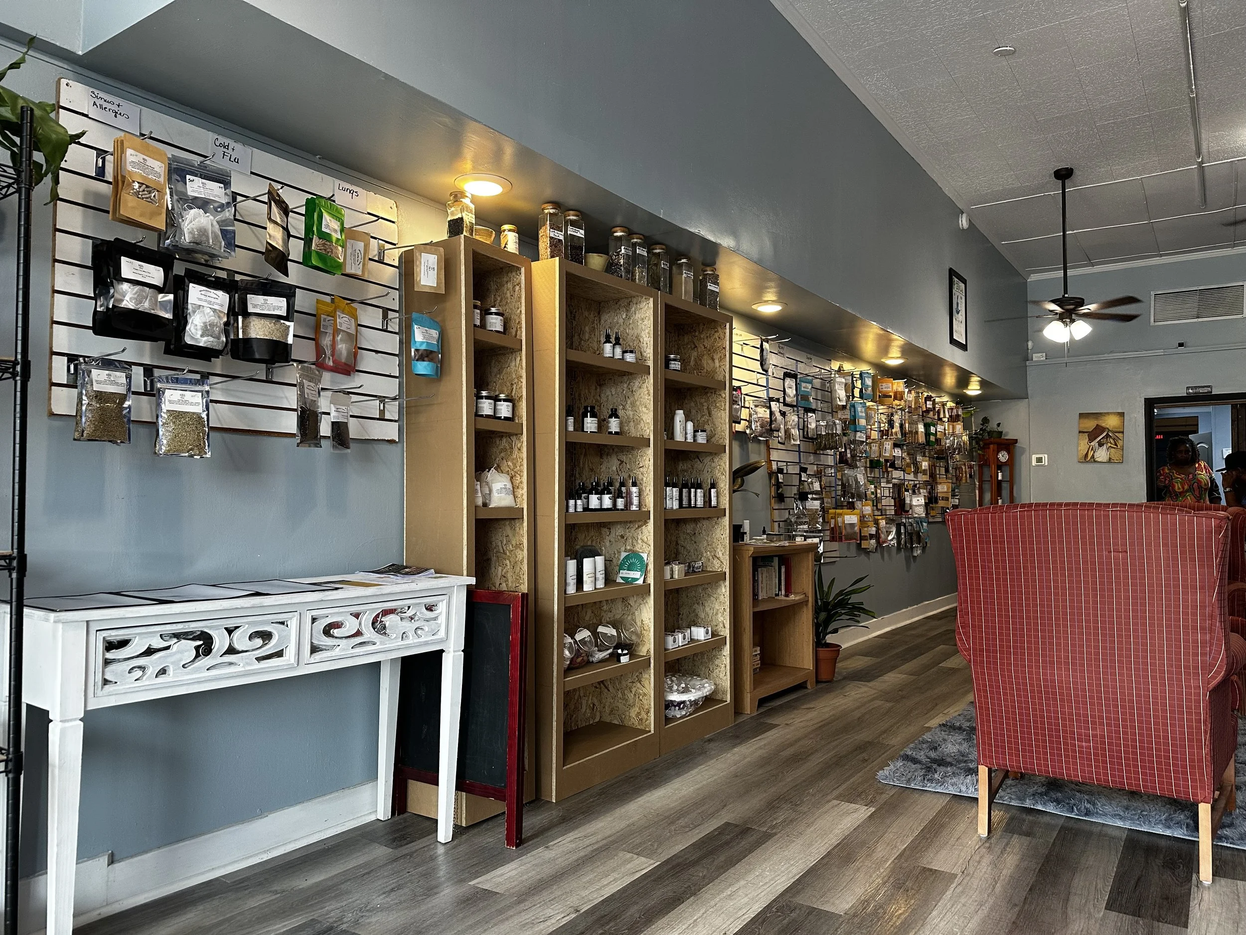 Interior of a store or cafe with wooden shelves filled with jars, bottles, and packages of herbs or spices. There is a white decorative table against a light blue wall with hanging packets of various herbs or spices. A red-checkered armchair is visible on the right, and a woman in colorful clothing is sitting in the background.