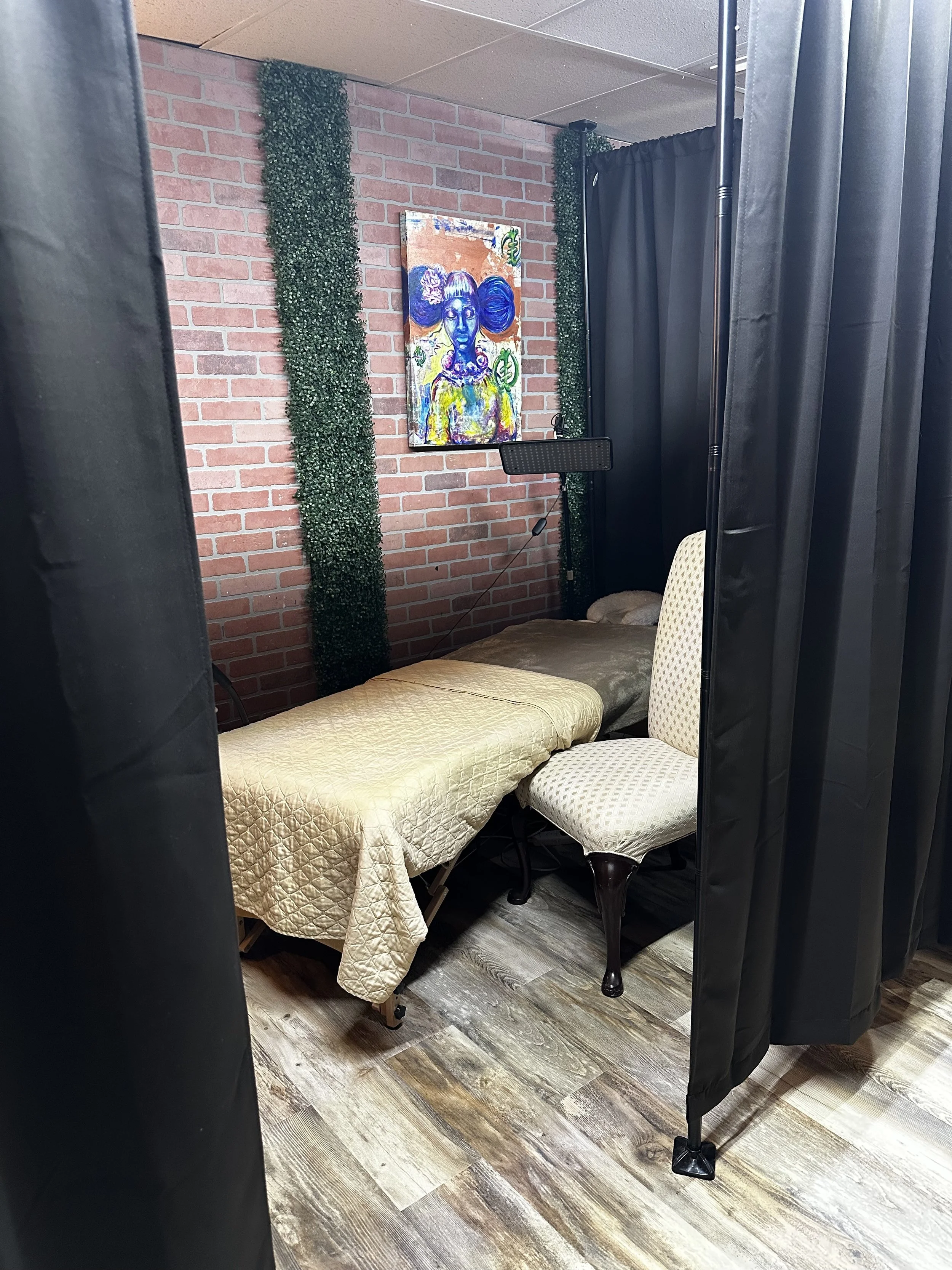 A staged room with a massage table covered with a quilted gold blanket, two chairs, a brick wall with decorative greenery, a colorful abstract portrait, and black curtains.