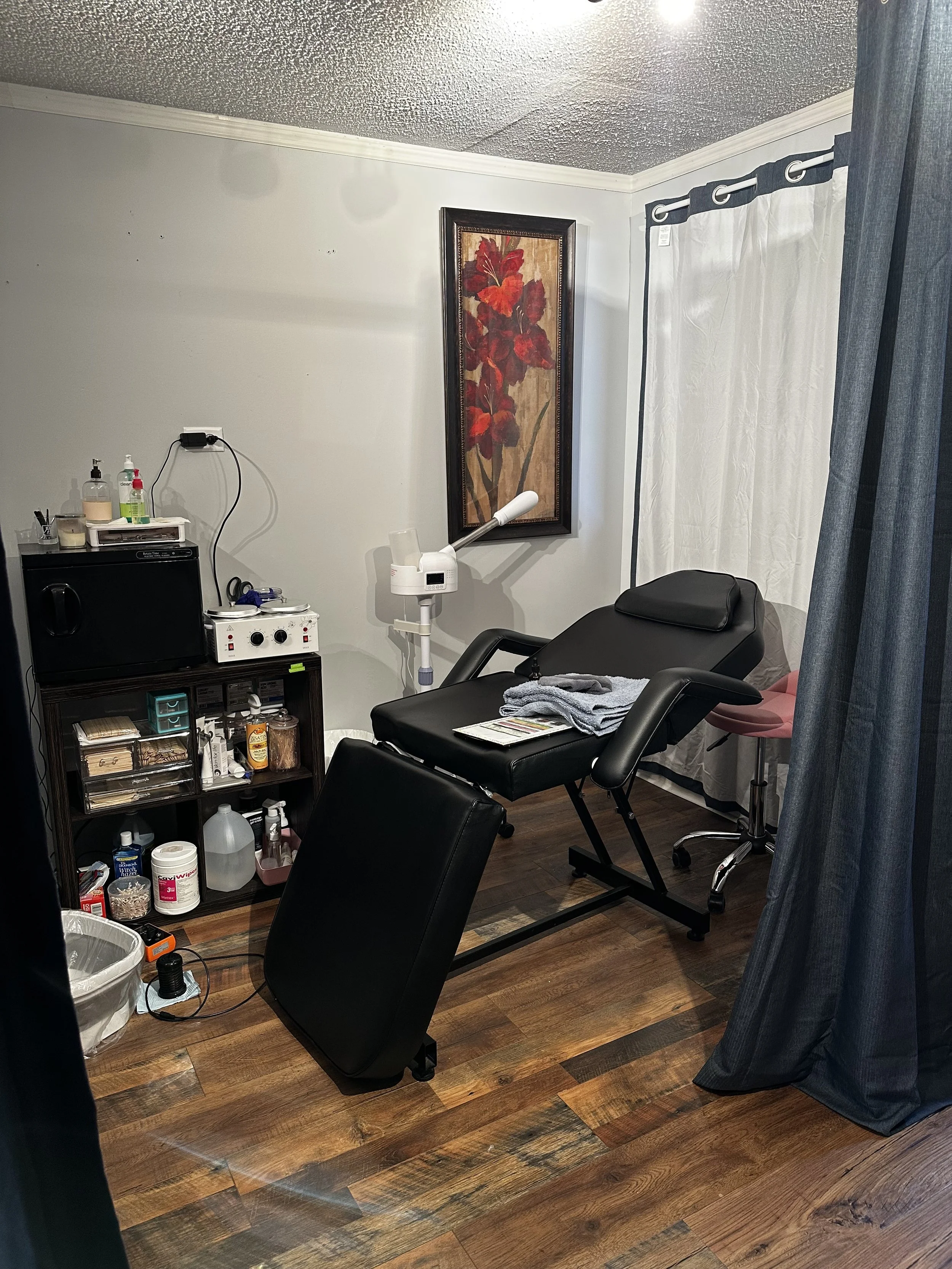 A medical or chiropractic examination room with a black reclining chair, a small cover with towels, a cabinet with supplies, a framed floral artwork on the wall, a white curtain, and various medical equipment including a lamp and machine.