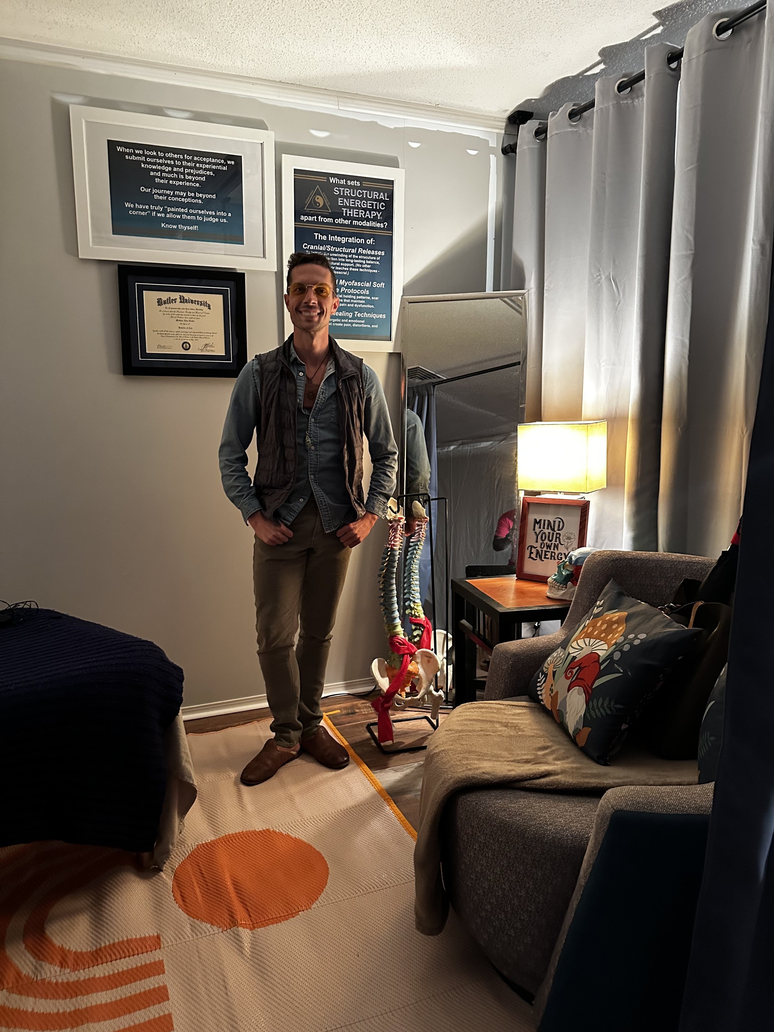 A man standing in a cozy living room, smiling at the camera. The room has various framed pictures and certificates on the wall, a colorful geometric rug on the floor, and a beige couch with patterned pillows. There is a lamp and a side table with decorative items, and a curtain covering a window or doorway.