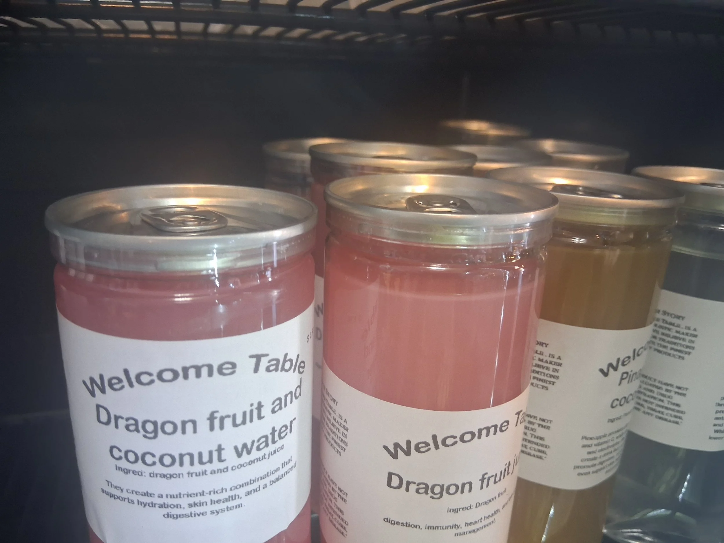 Close-up of canned fruit beverages with labels reading "Welcome Table" and featuring ingredients like dragon fruit, fig, and coconut water.