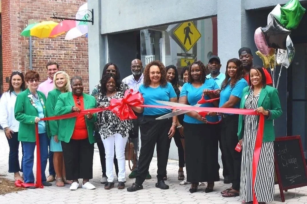 Group of people at a ribbon-cutting ceremony outside a storefront, with colorful balloons and balloons shaped like stars, some holding scissors, celebrating a grand opening.