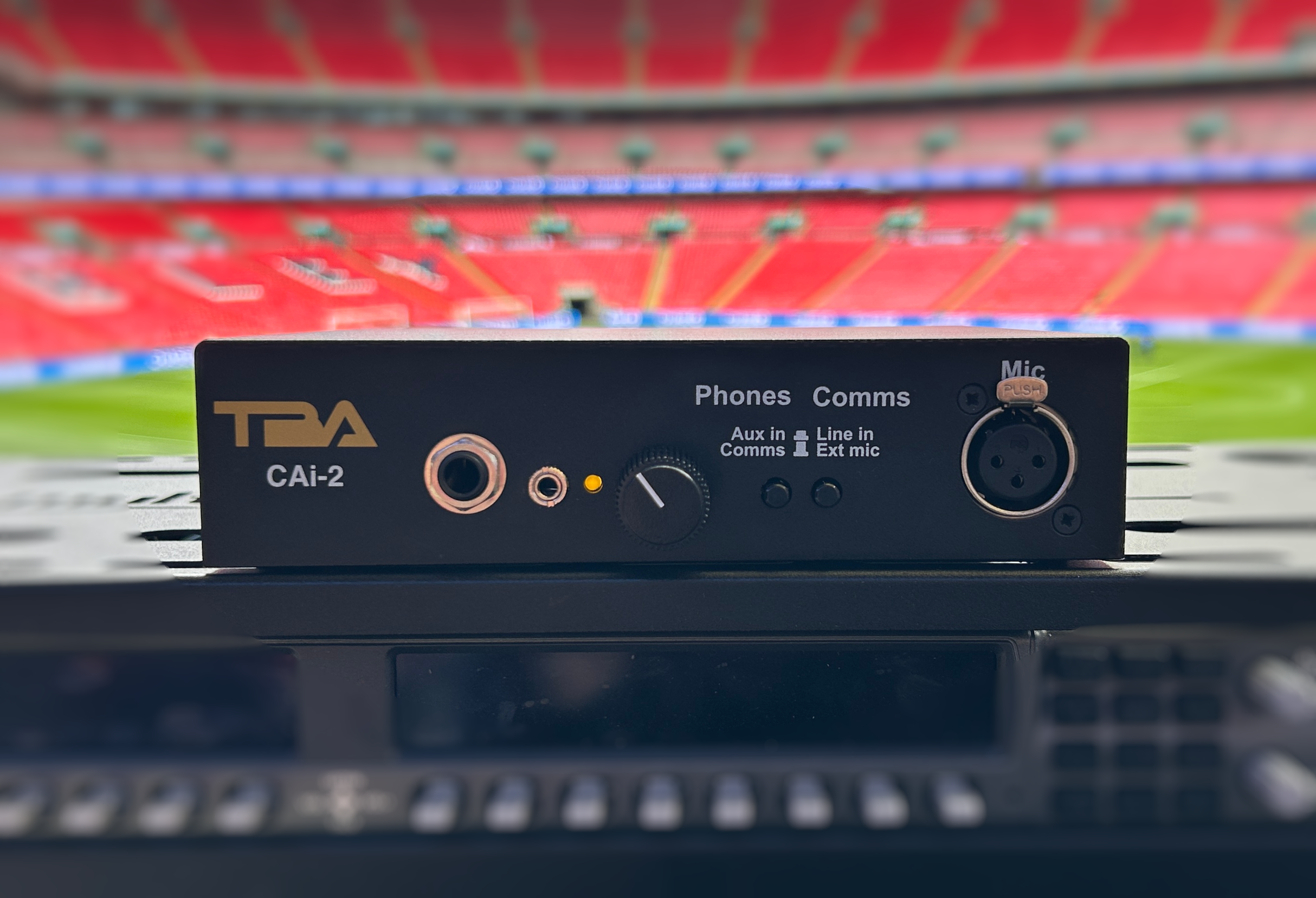 Close-up of a black audio interface labeled 'TDA CAi-2' with various input ports and controls, with blurred red stadium seating in the background.