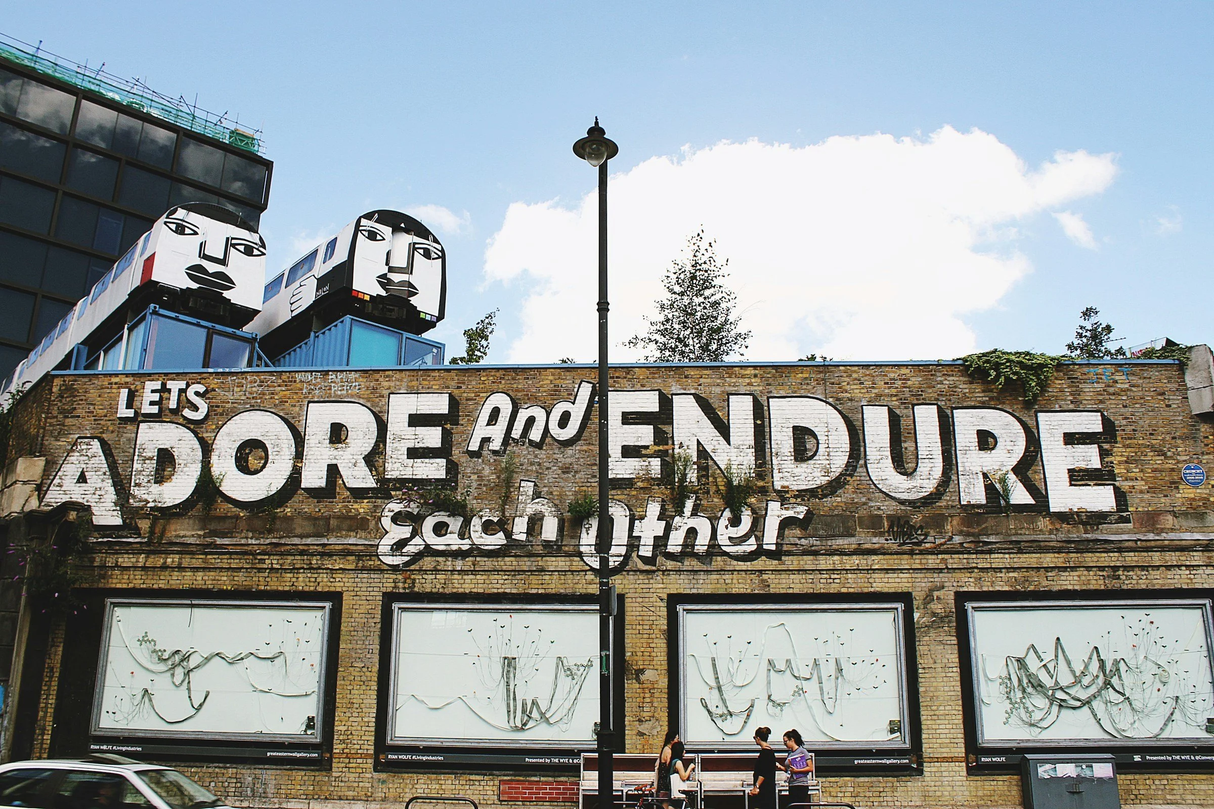 A wide shot of a brick building, he top of the wall displays the message "LETS ADORE AND ENDURE EACH OTHER" in bold white paint.On the roof, two decommissioned London Underground train carriages
