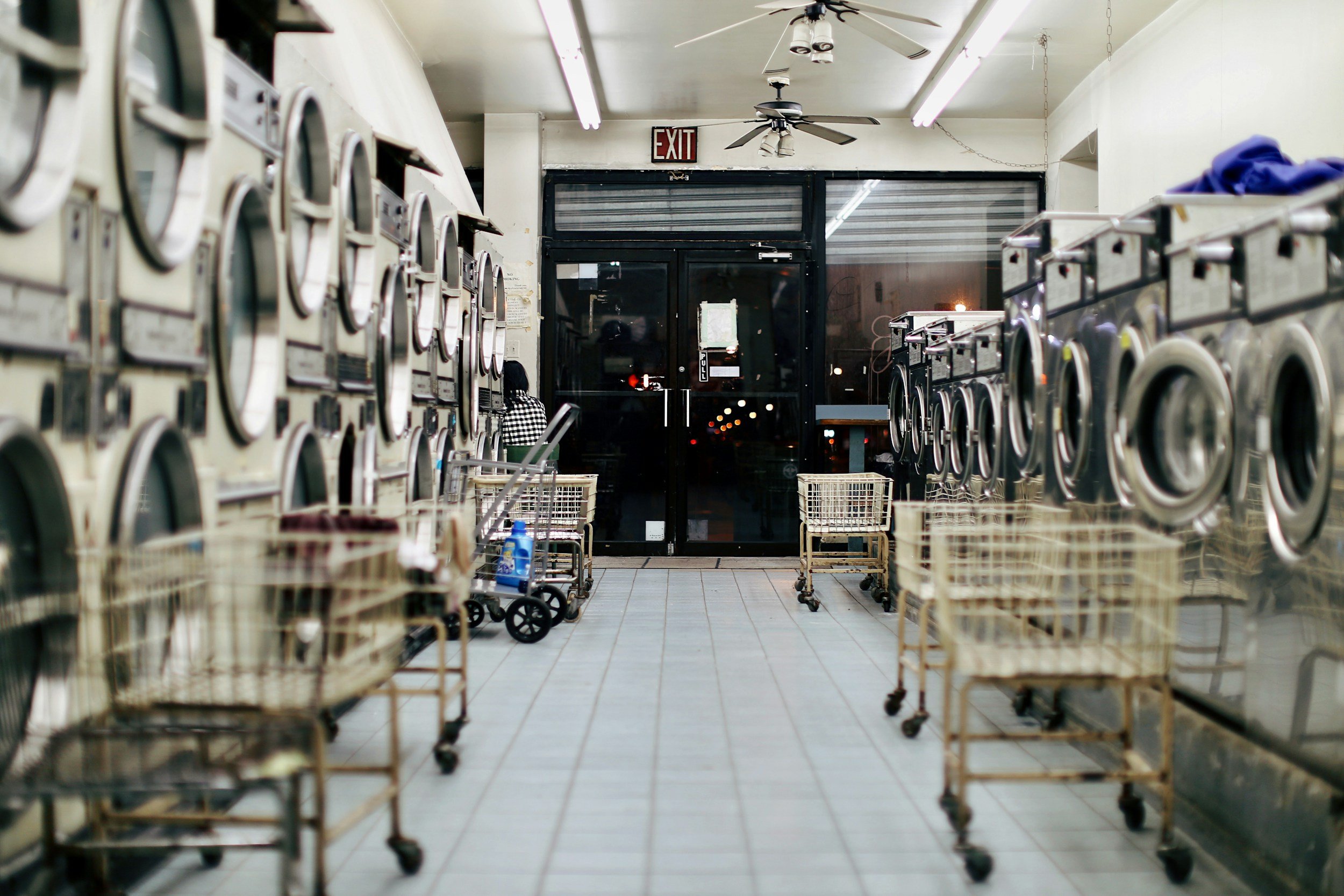 Empty laundromat with stacked washing machines, laundry carts, and a person sitting at the back near the entrance door.
