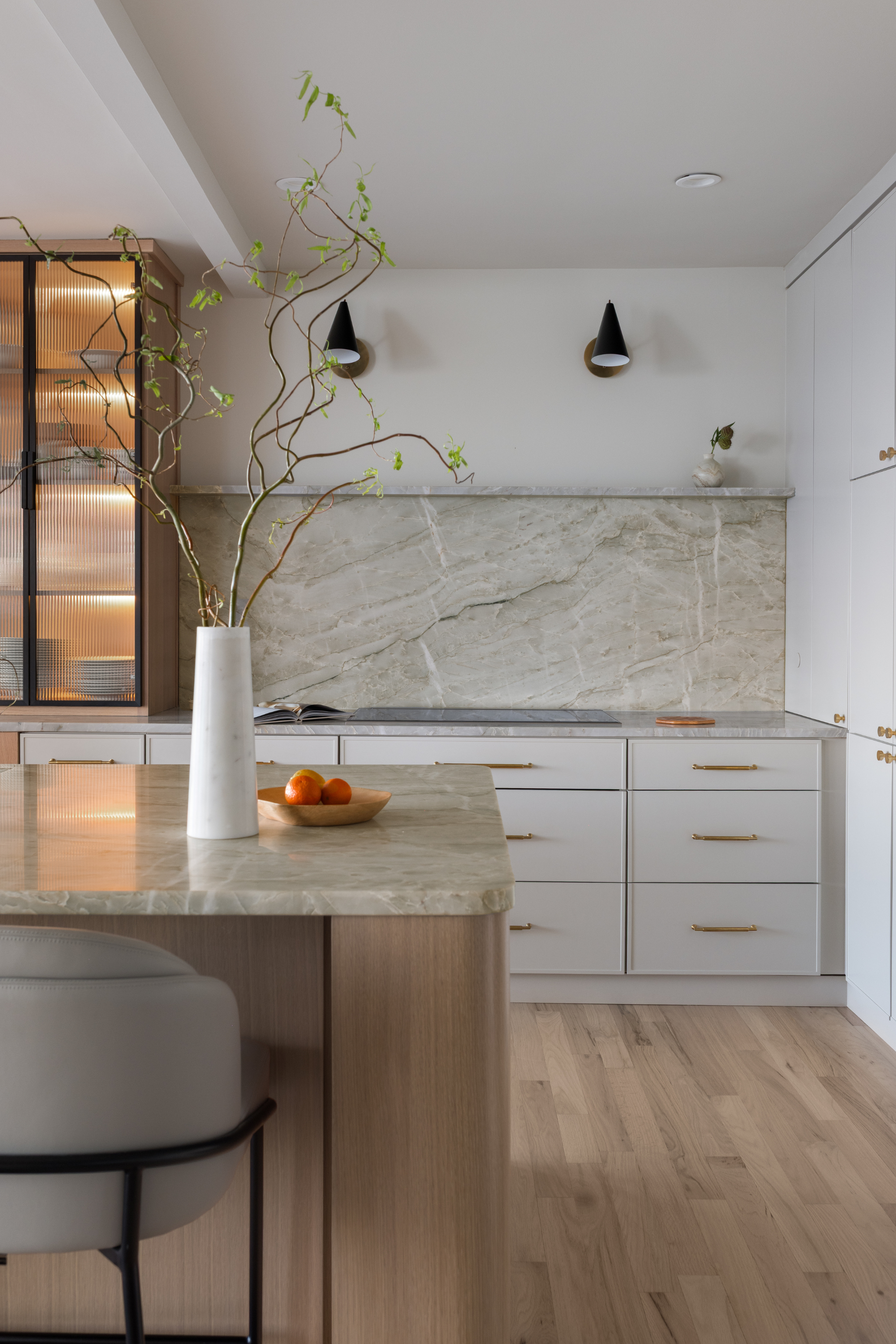 Modern kitchen with a marble countertop, white cabinets, and a wooden floor. There's a white vase with a twisting plant and a bowl of oranges on the counter. Black wall sconces and a small white decorative object are on the wall.