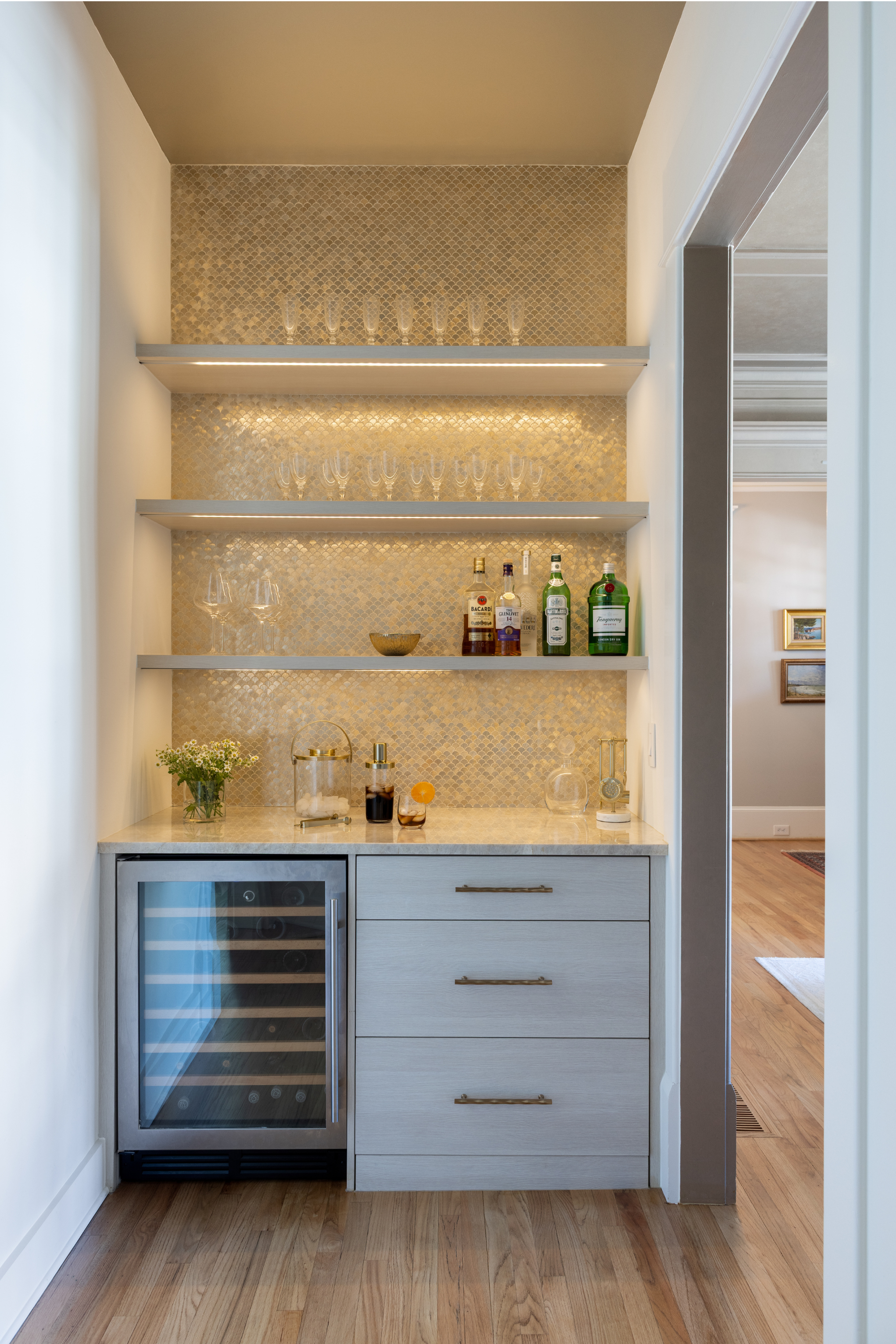 A built-in bar area with three shelves holding glasses and liquor bottles, a marble countertop, a small wine fridge, and decor items, with a wall featuring gold mosaic tiles behind.
