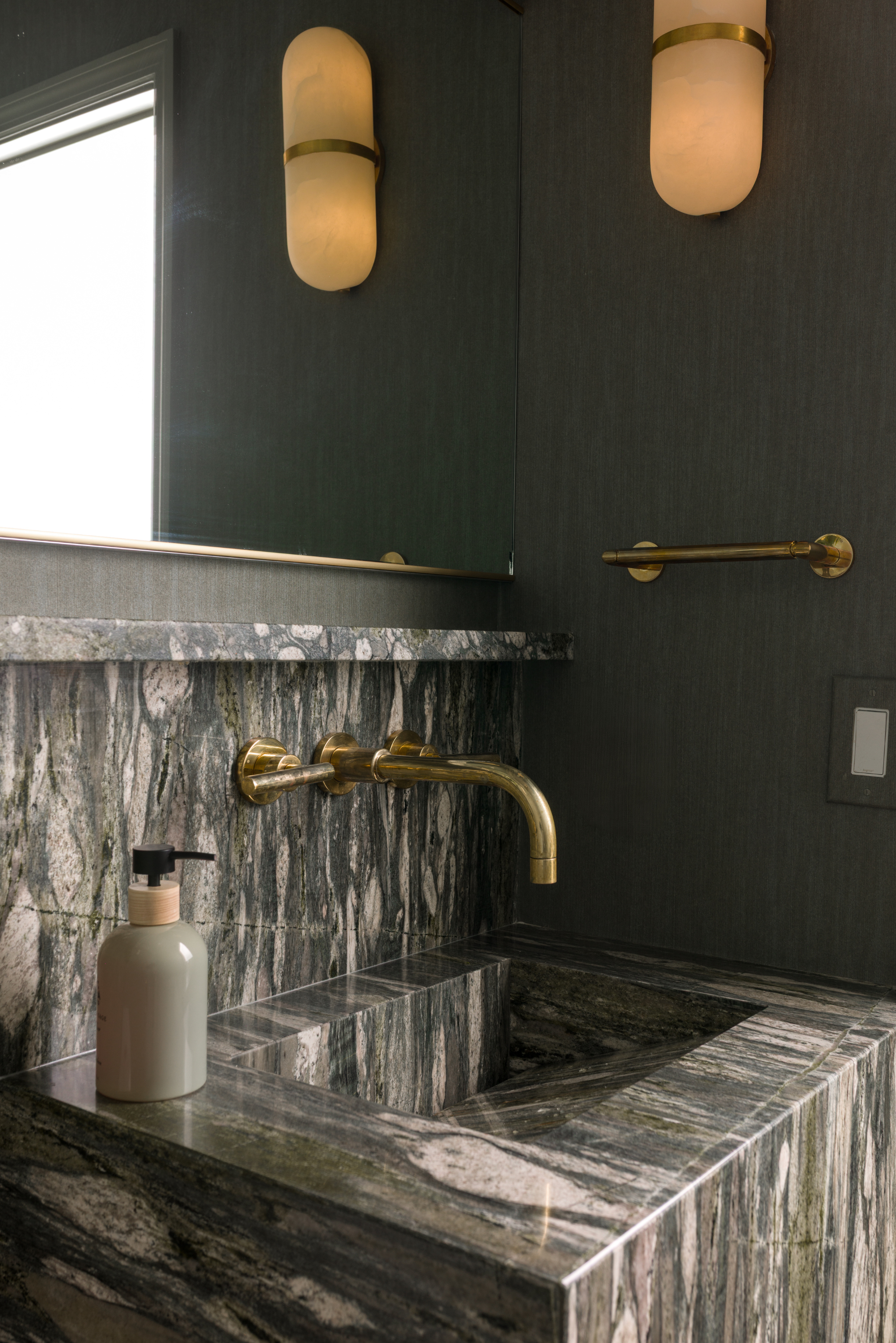 Bathroom sink with a marble countertop and brass fixtures, a wall-mounted bottle of liquid soap, a mirror, and modern wall sconces with frosted glass shades.
