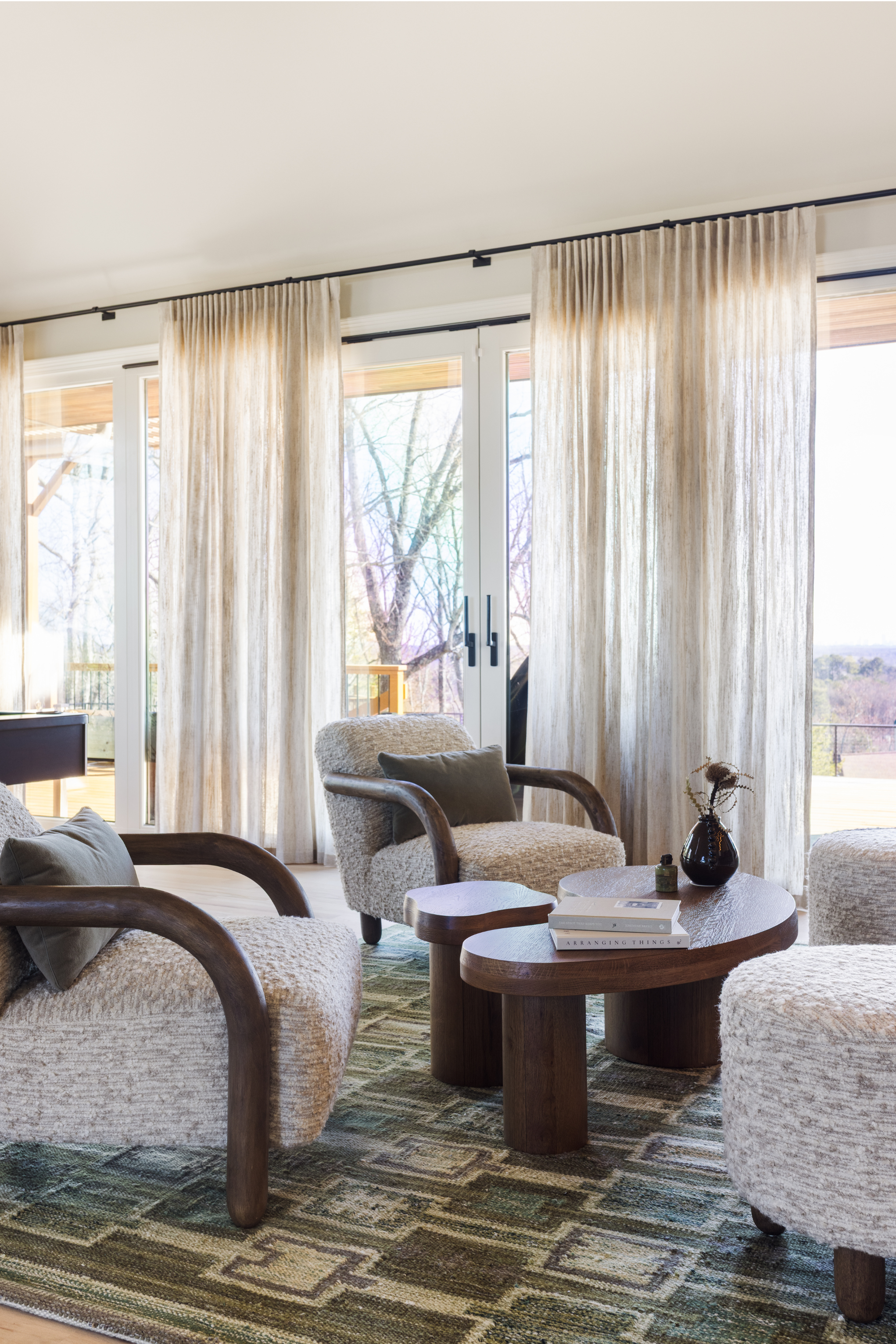 Living room with large sliding glass doors, beige curtains, cozy armchairs with wooden arms, a wooden coffee table with books and a vase, and a patterned rug. Outside, trees are visible through the glass doors.