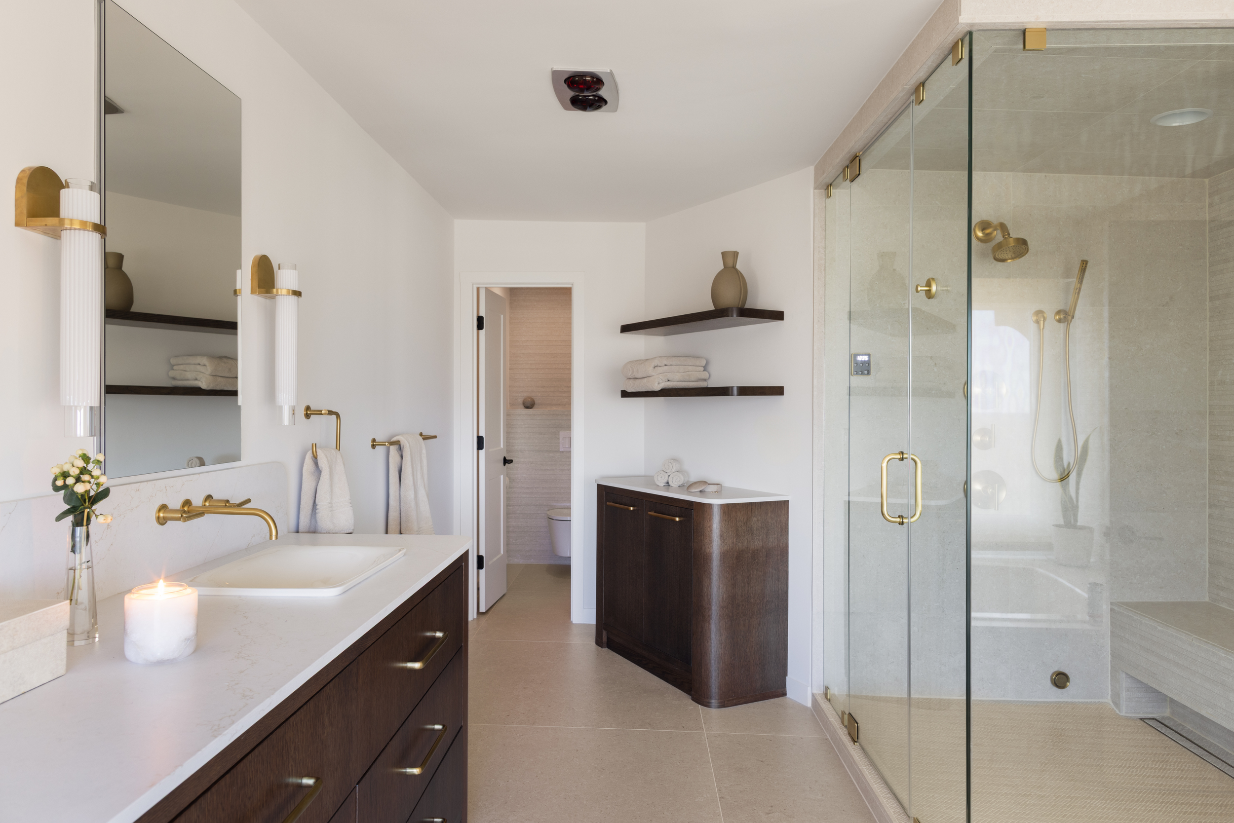 Modern bathroom with dark wood vanity, white countertop, large mirror, glass shower enclosure with gold fixtures, beige flooring, wall-mounted shelves with towels and vases, and a doorway leading to a toilet room.