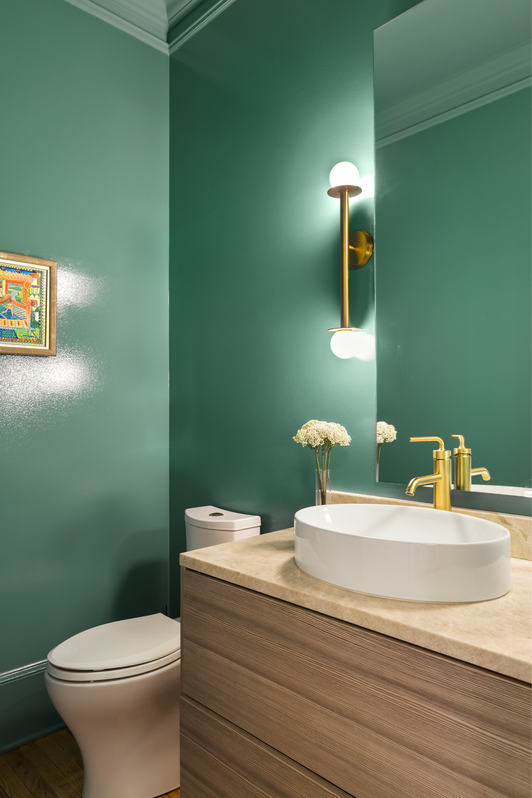 Modern bathroom with green walls, a white toilet, a beige marble countertop with an oval vessel sink, a large mirror, and a wall-mounted gold faucet. A small vase with white flowers is on the counter, and a gold wall sconce with two spherical lights is mounted above the mirror.
