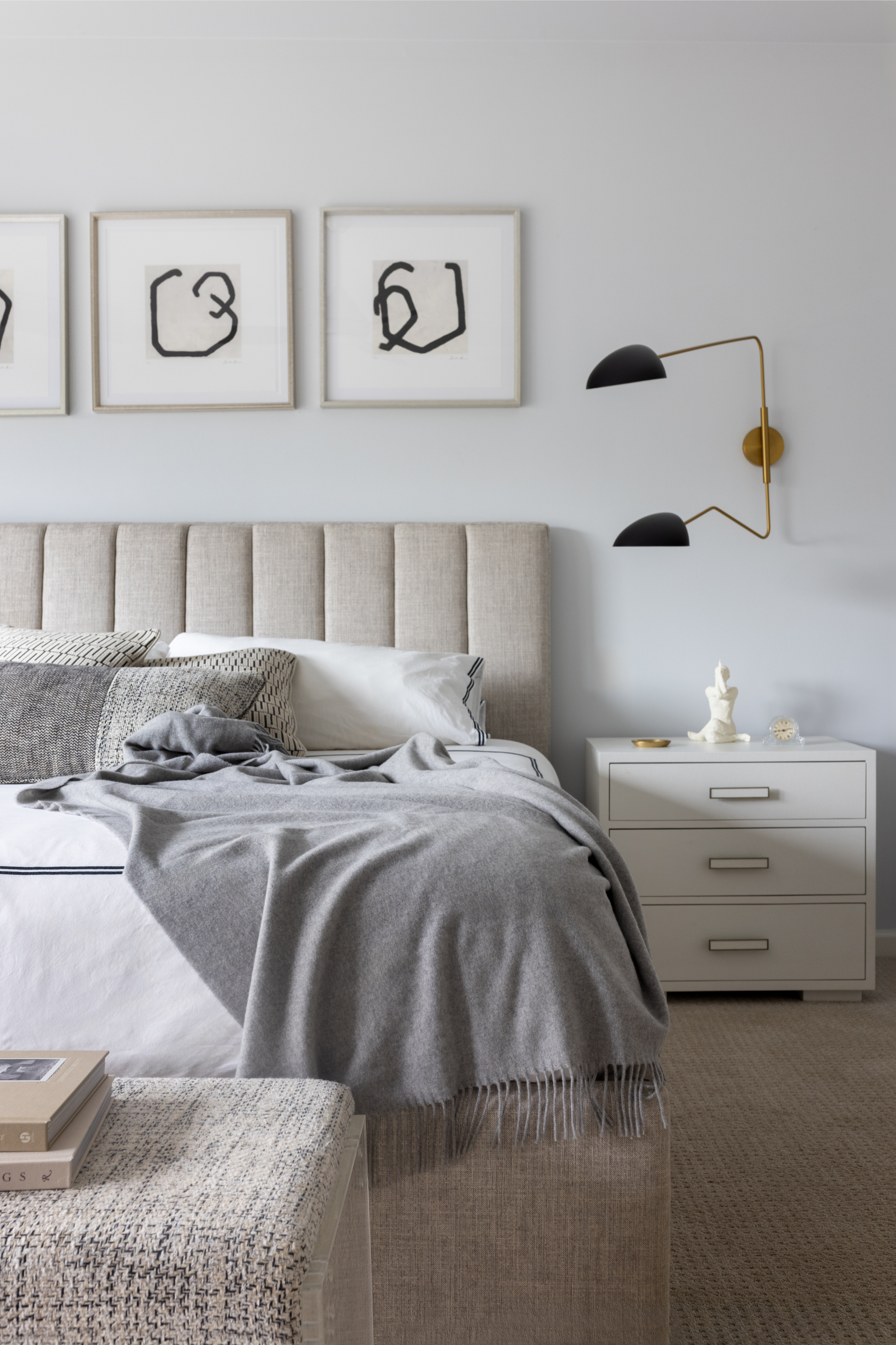 A cozy bedroom with a beige upholstered headboard, white bedding, and a gray throw blanket. Artworks with abstract black scribbles hang on the light gray wall above the bed. A white nightstand with decorative items, including a small sculpture and clock, is next to the bed. A modern black and gold wall sconce provides lighting.