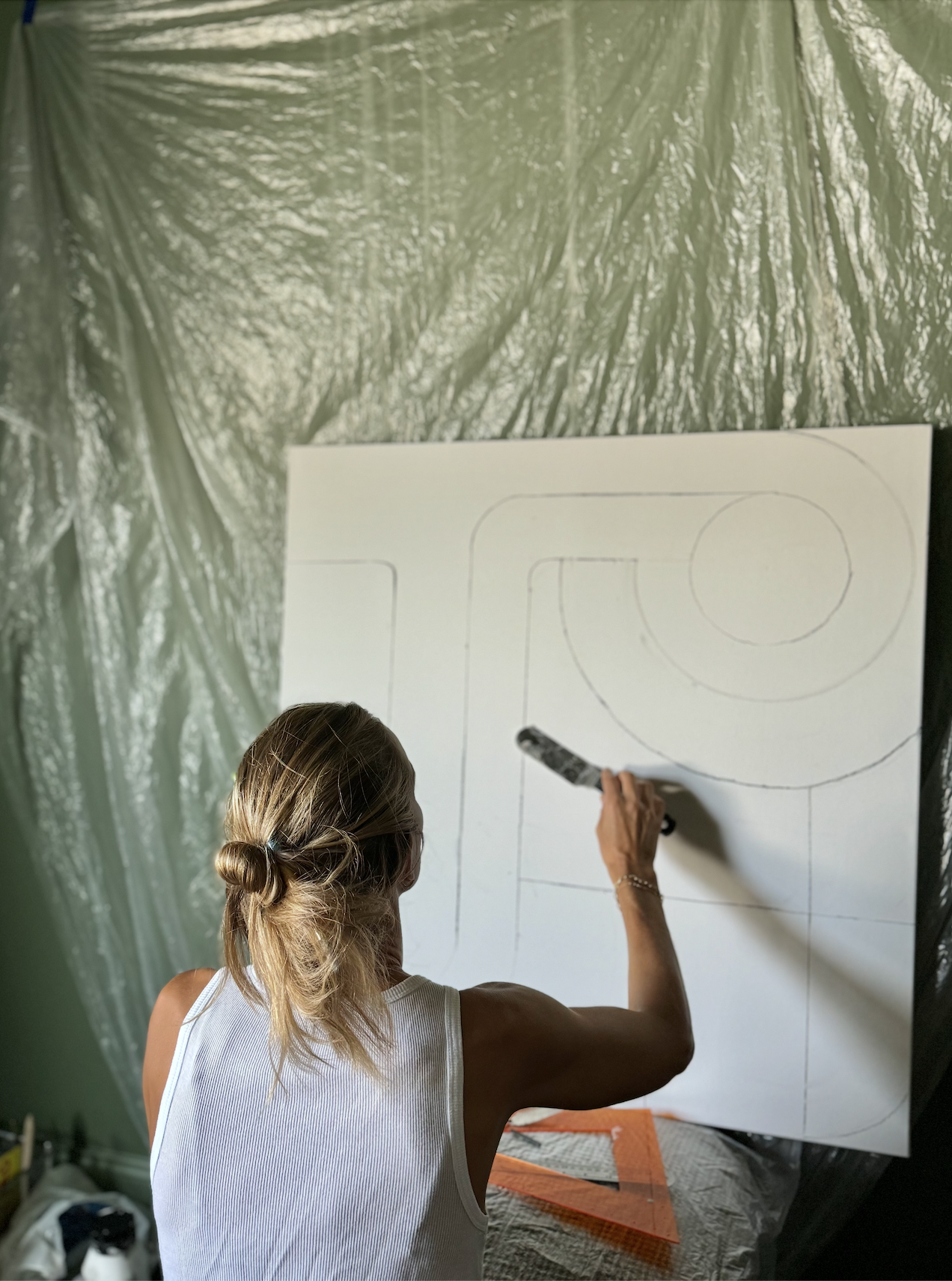 A woman with blonde hair tied in a bun, wearing a white sleeveless top, is drawing geometric shapes on a large white canvas with a marker, in a room covered with plastic sheeting.