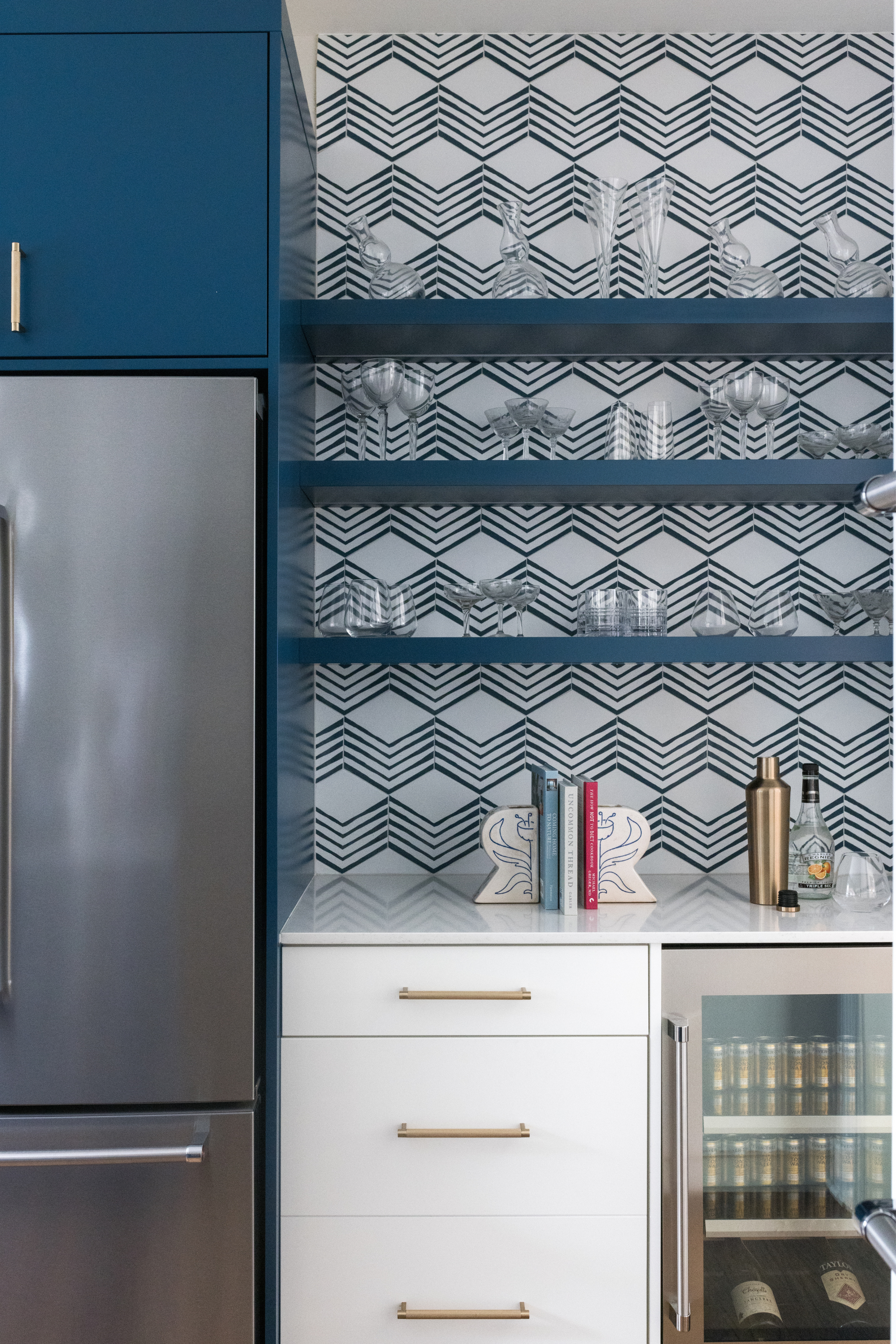 Kitchen cabinet with open blue shelves displaying glassware, patterned wallpaper backdrop, and a white countertop with books and decorative items.