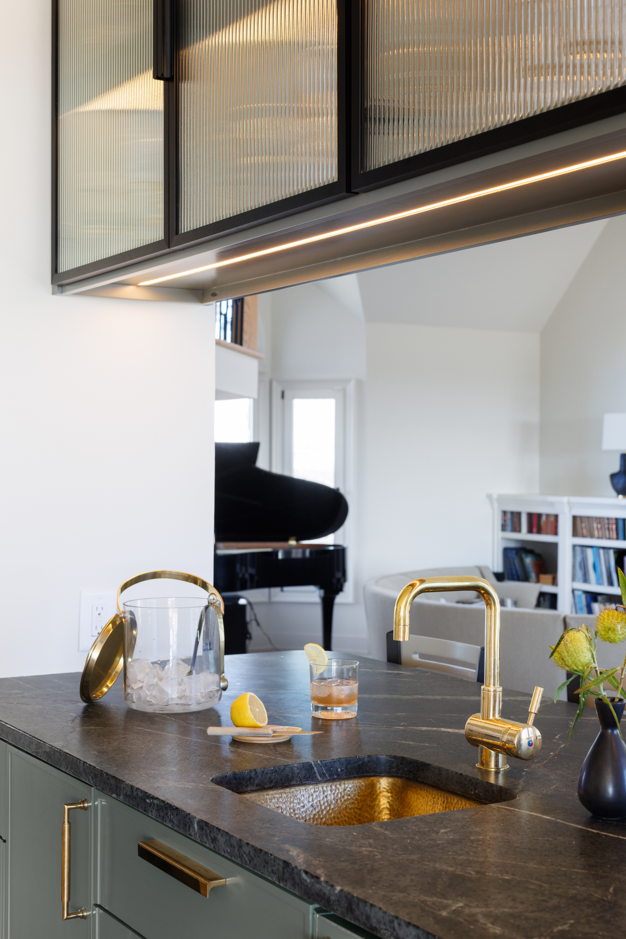 Modern kitchen with dark stone countertop, gold sink faucet, and transparent ice bucket with drinks on the island. In the background, there is a living room with a black grand piano, bookshelf, and white walls, illuminated by natural light through windows.