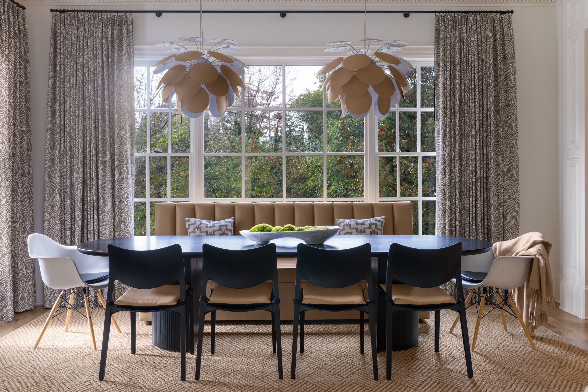 Modern dining room with large window, black oval table, white and black chairs, beige bench with throw pillows, two pendant lights, patterned curtains, and a bowl of green moss in the center.