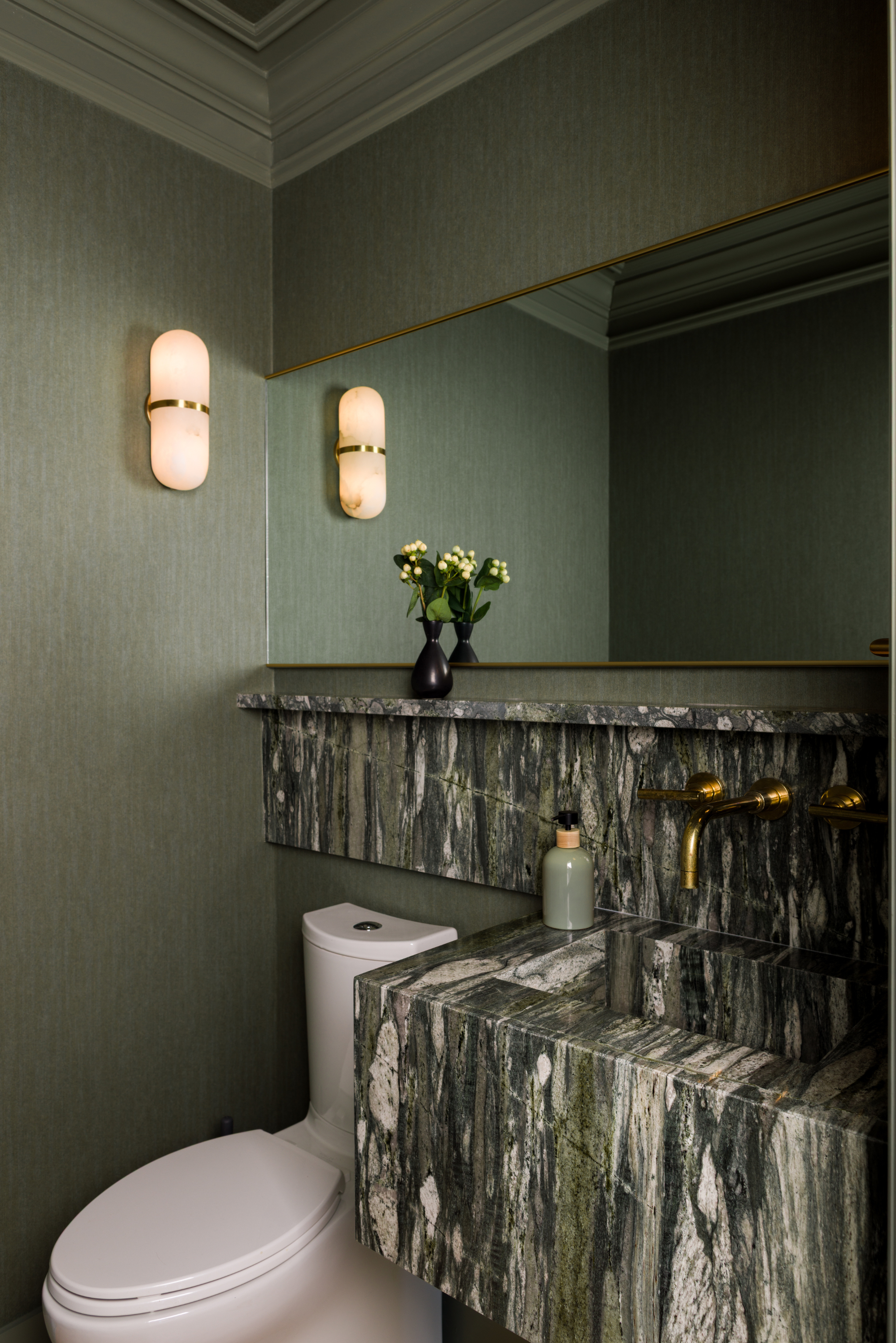 A bathroom with green textured walls, gold wall sconces, a large mirror, a marble sink with a gold faucet, a vase with flowers, and a white toilet.