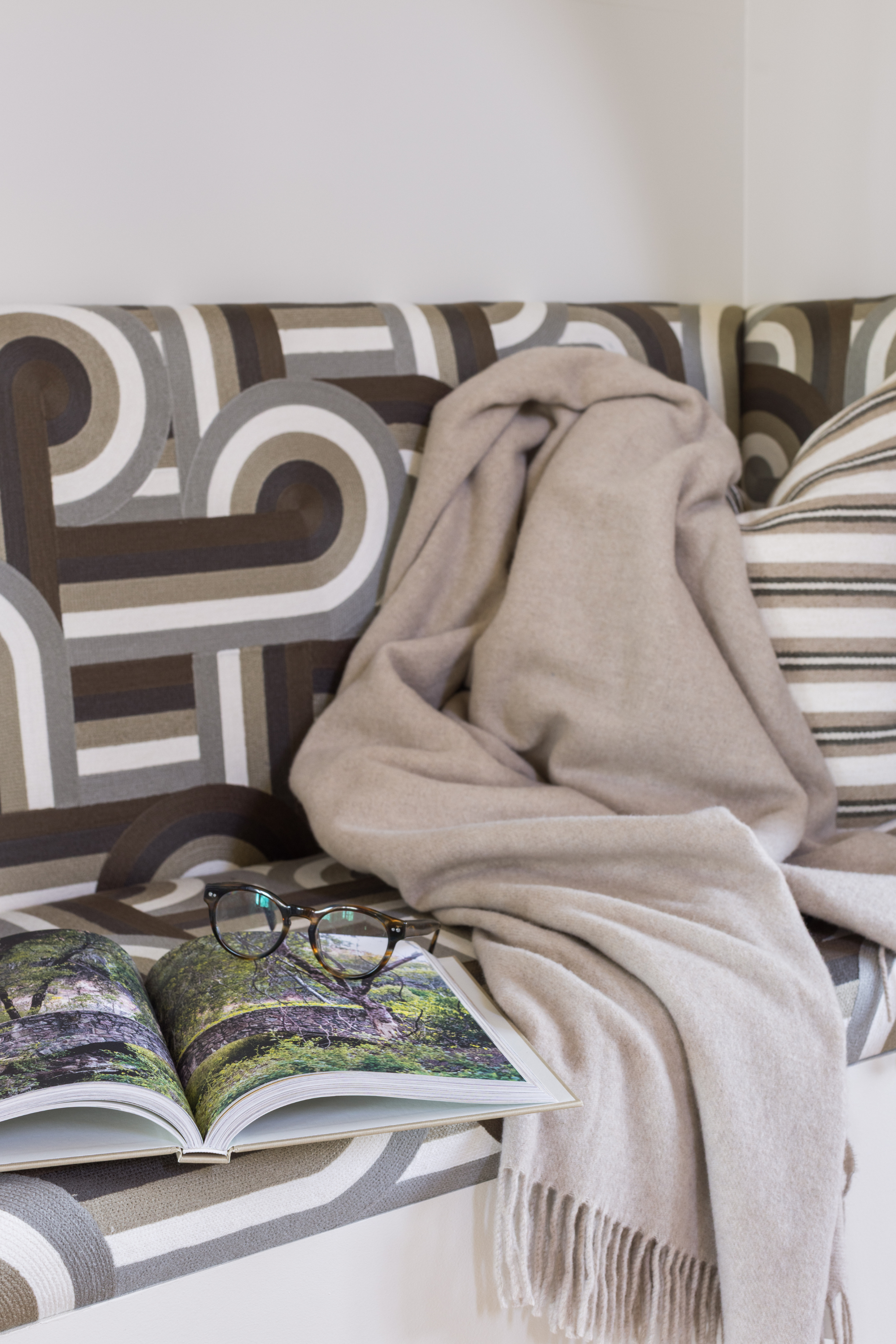 A cozy living room corner with a patterned sofa, a beige throw blanket, an open book with scenic photos, and a pair of eyeglasses on the book.