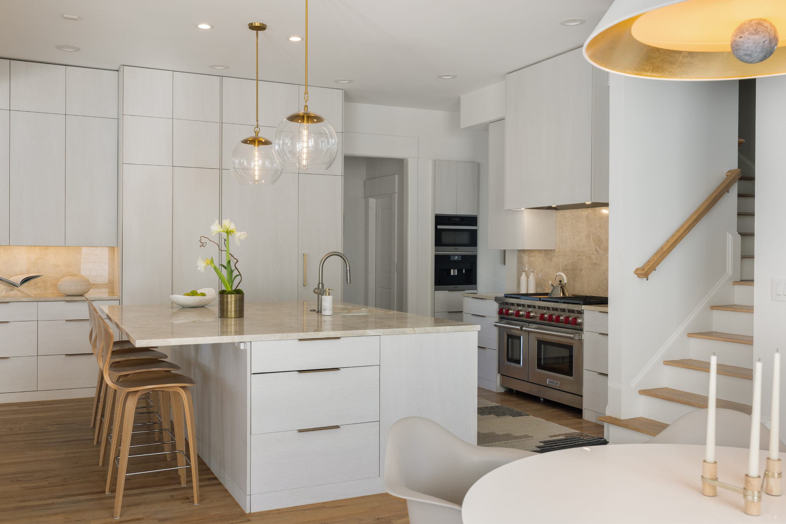 Modern kitchen with white cabinetry, marble island, and wooden bar stools. Features a stainless steel stove, gold pendant lights, and a staircase nearby.