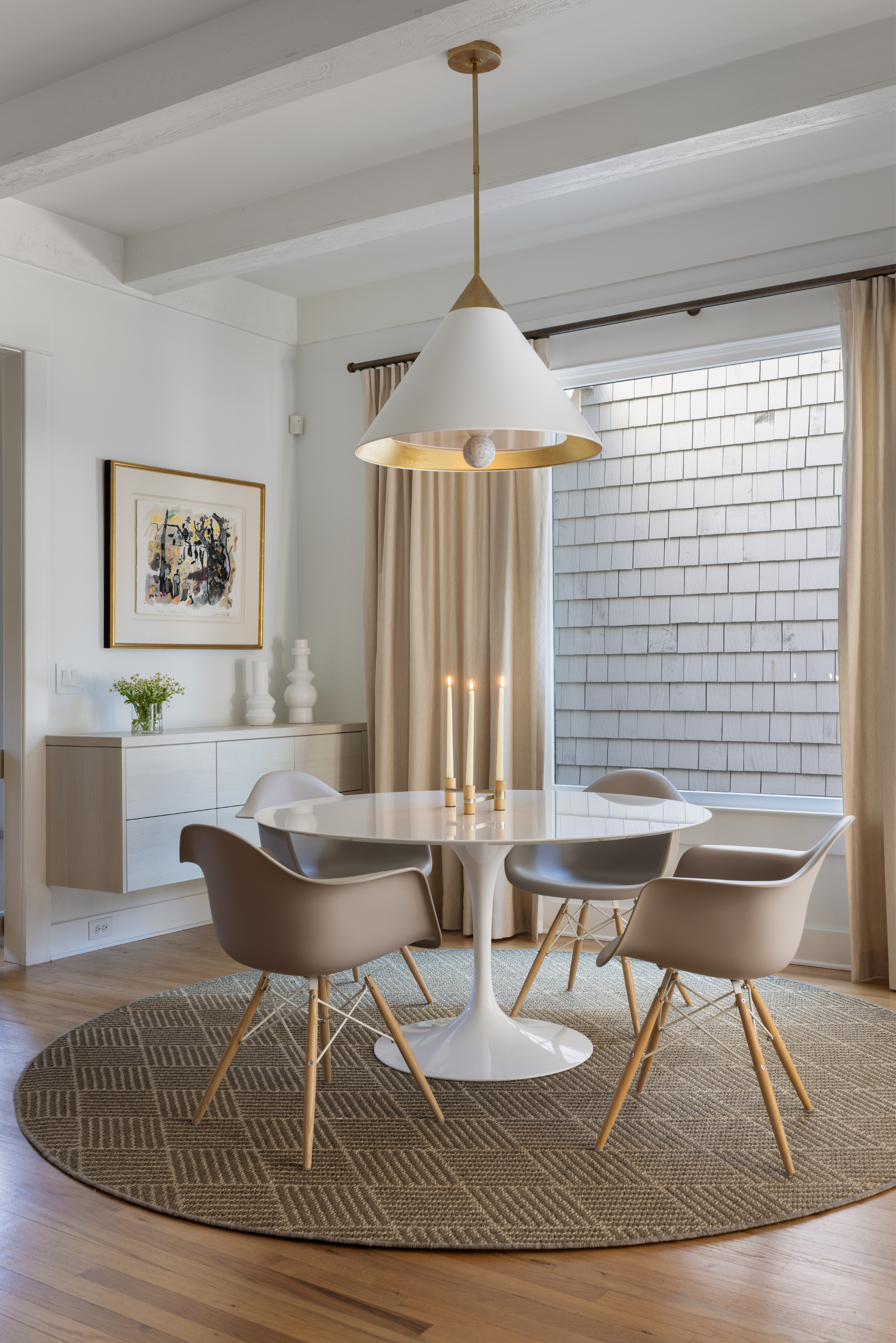 Dining room with a white round table, four beige chairs, a modern pendant light, a beige curtain, and a window with shingled exterior wall, decorated with candles and artwork on the wall.