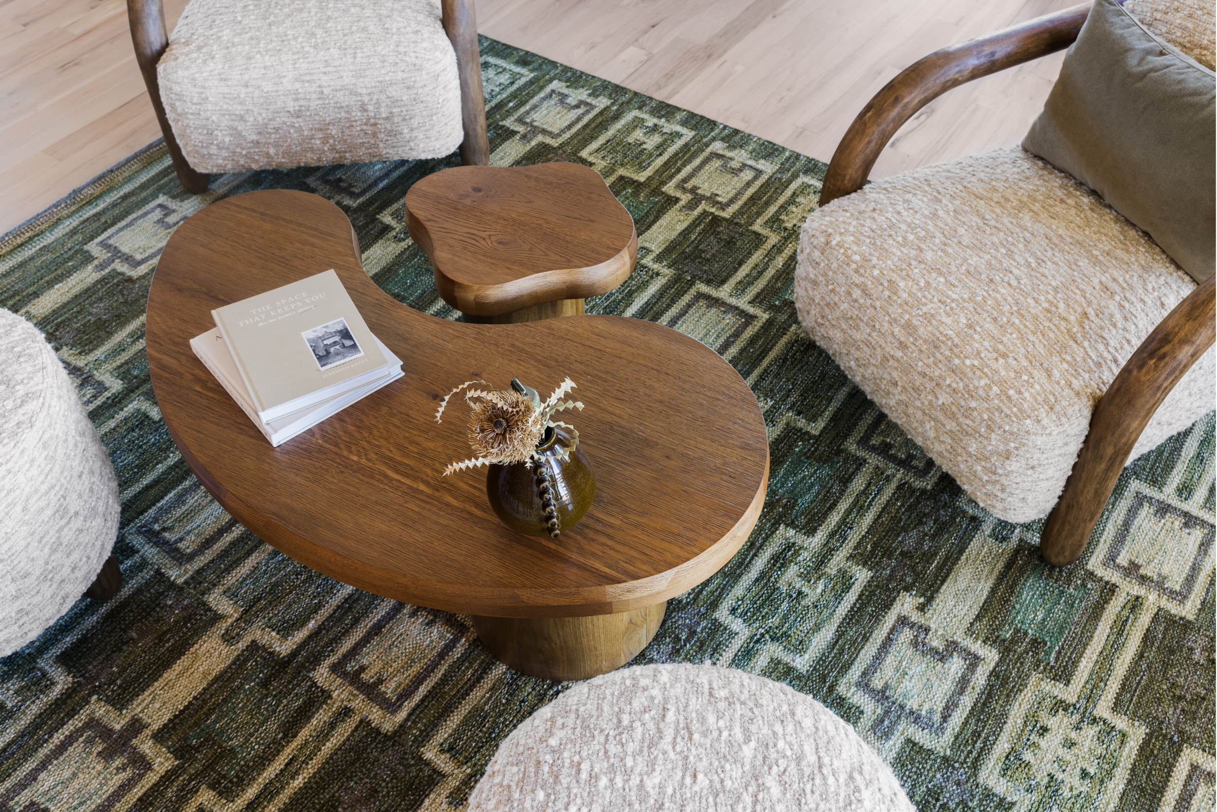 A cozy living room corner featuring a wooden coffee table with a book and a vase with dried flowers, surrounded by upholstered chairs on a patterned rug.