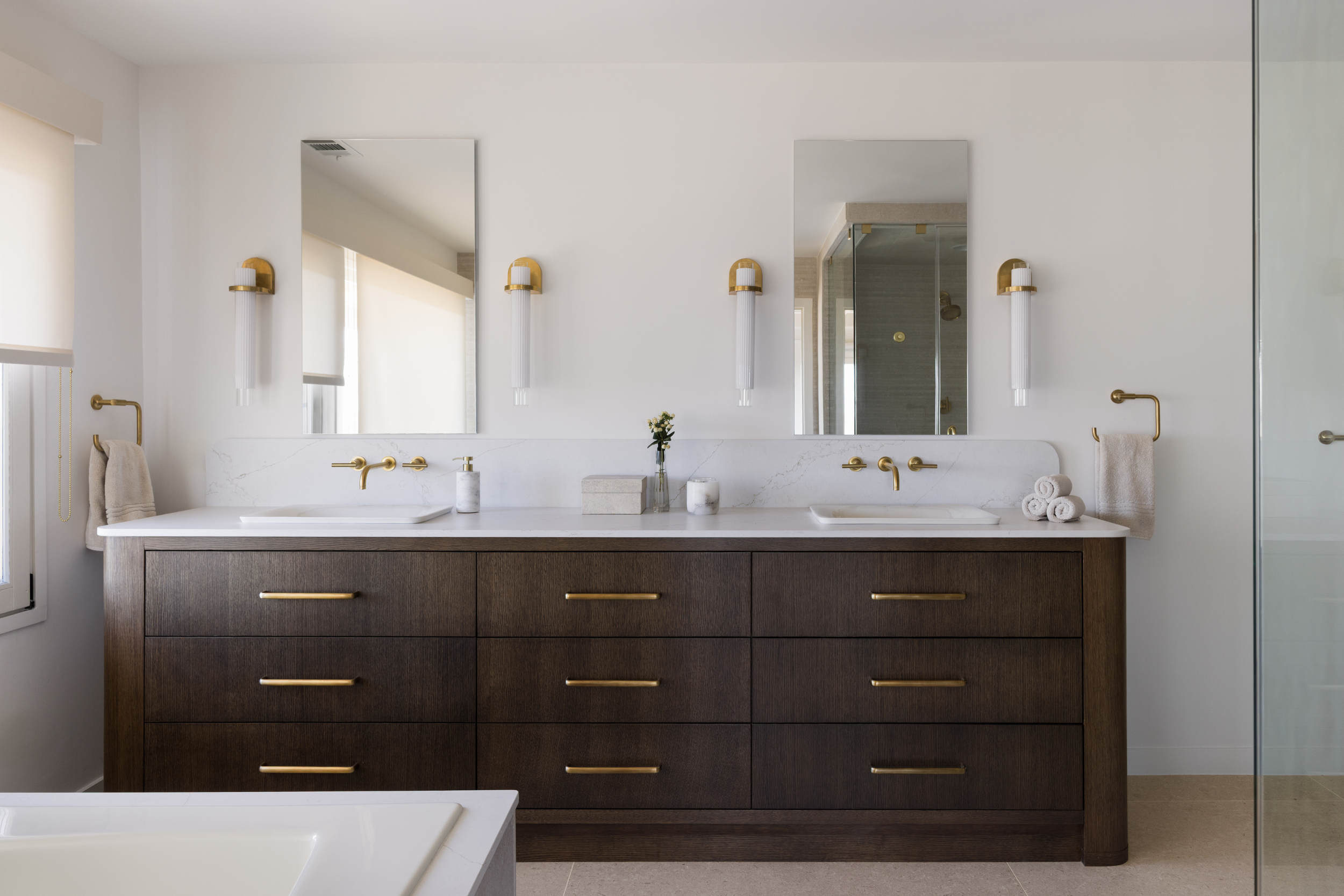 Modern bathroom with double vanity, white countertops, gold fixtures, mirrors, wall-mounted lights, towels, and a glass shower enclosure.